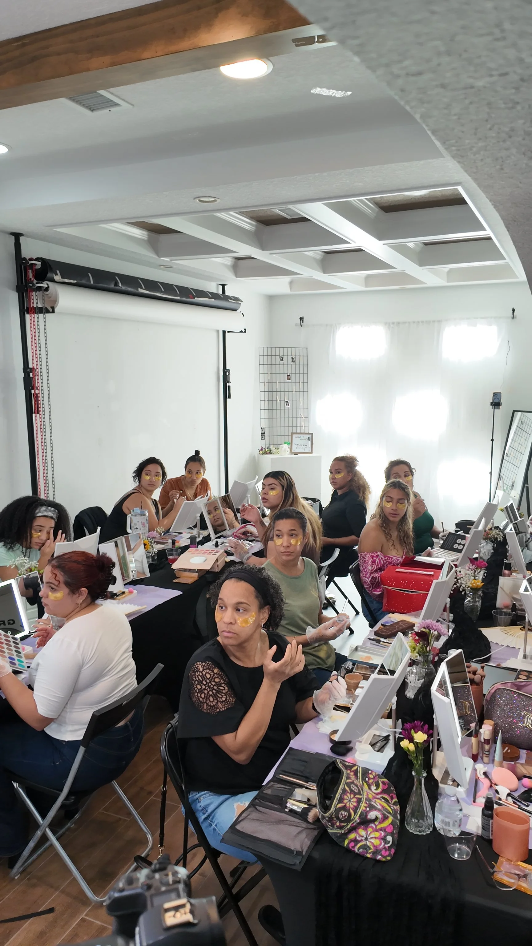 A group of women sitting at tables in a bright room, preparing makeup, with mirrors, makeup supplies, and decorative flowers on the tables.