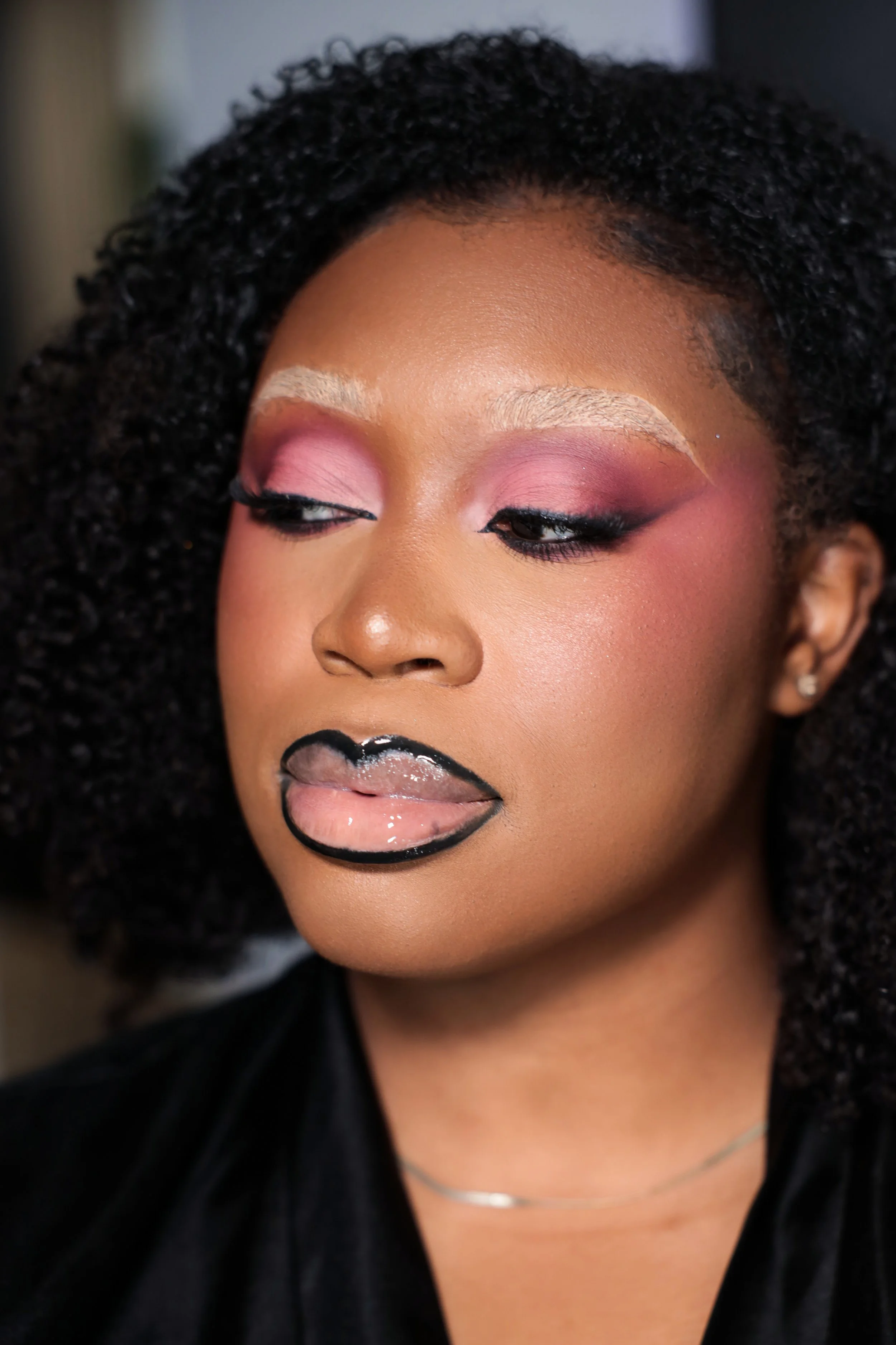Close-up of a woman with black curly hair, makeup including pink eyeshadow, black eyeliner, and glossy lipstick with a black outline. She has a light complexion and wears a silver necklace.