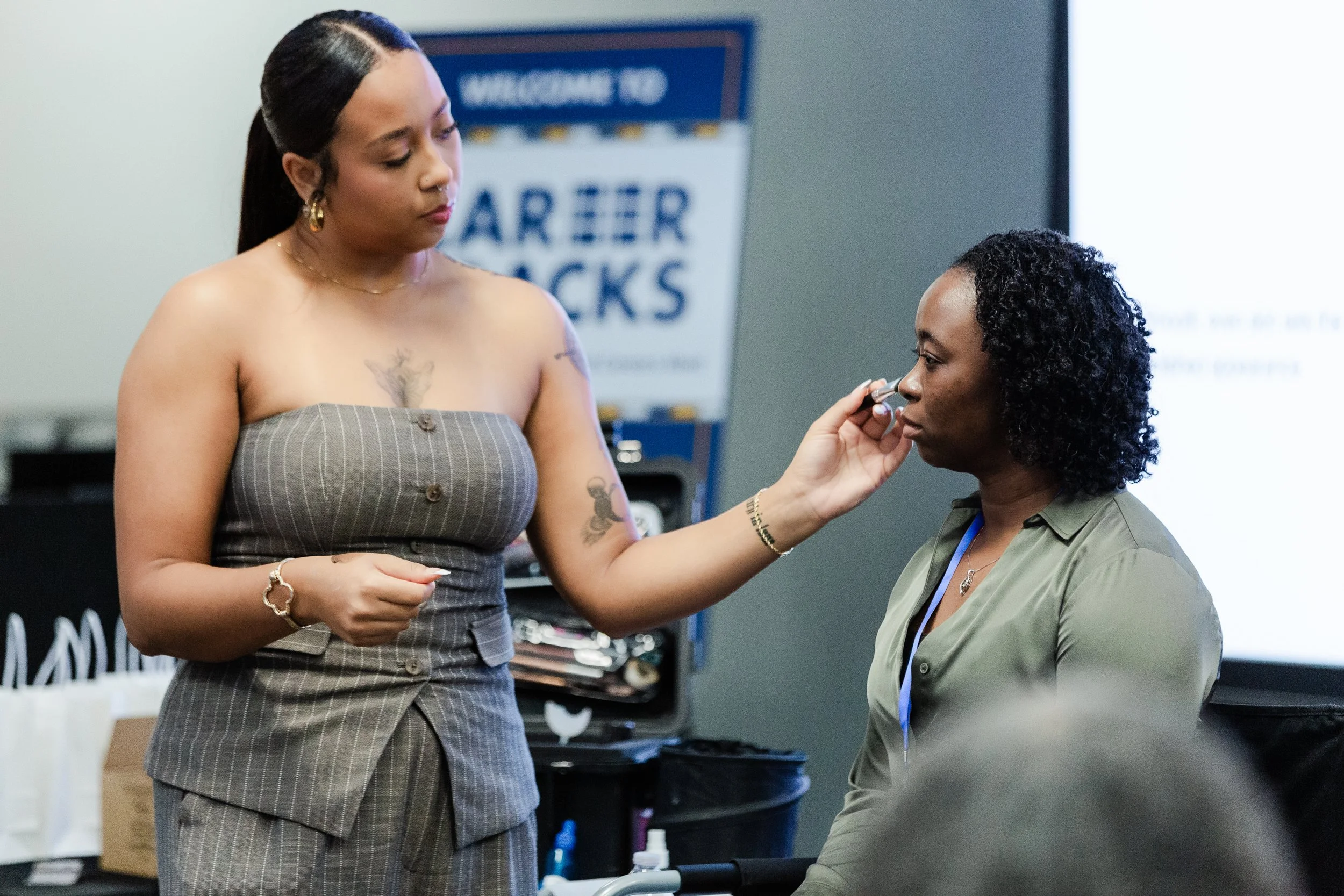 A woman with long dark hair and tattoos on her chest is applying makeup or skincare to another woman with curly short hair seated. Wounded warriors project.