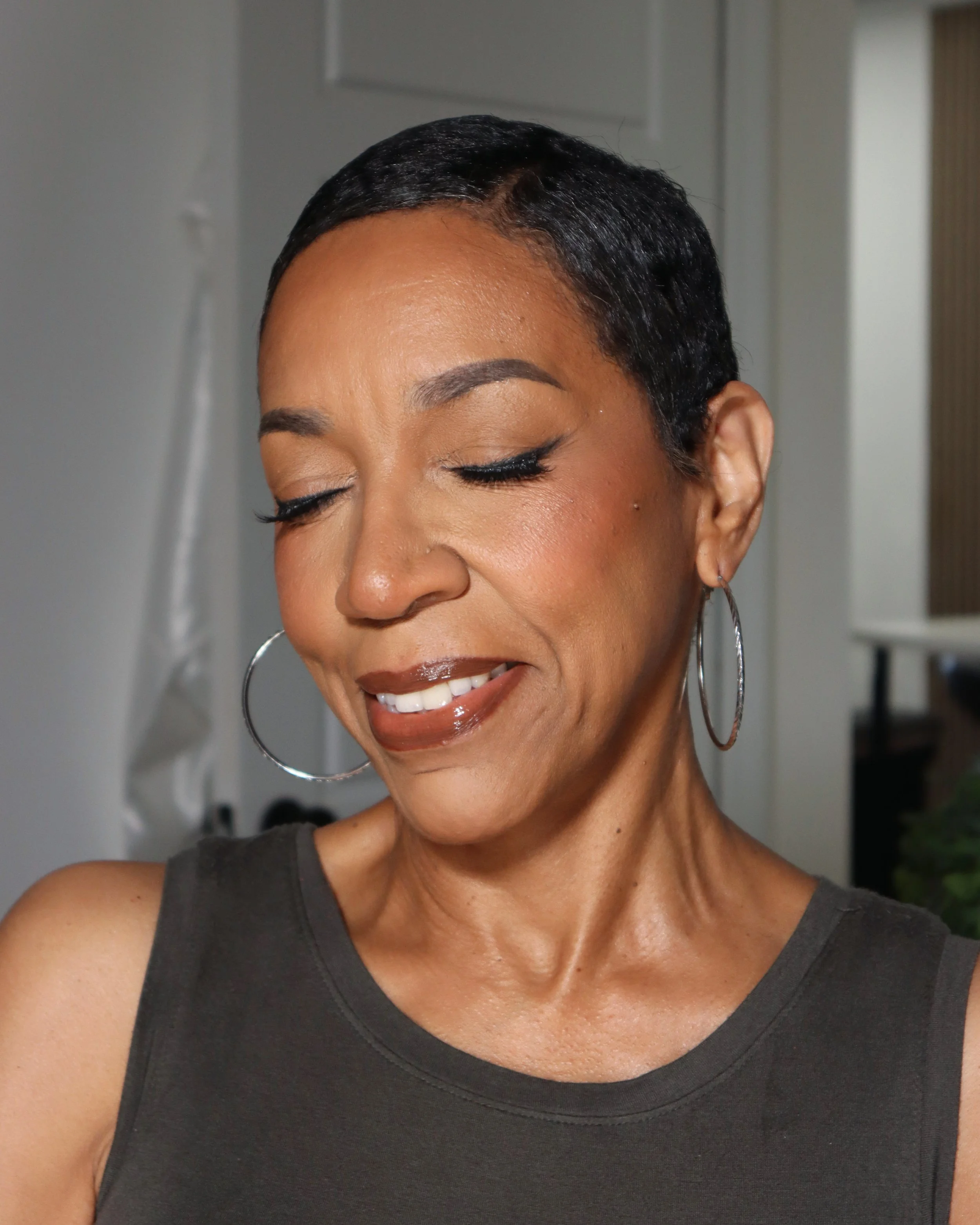Close-up of a woman with closed eyes, wearing hoop earrings, smiling, in a sleeveless dark top, with short dark hair, indoors with a blank background.