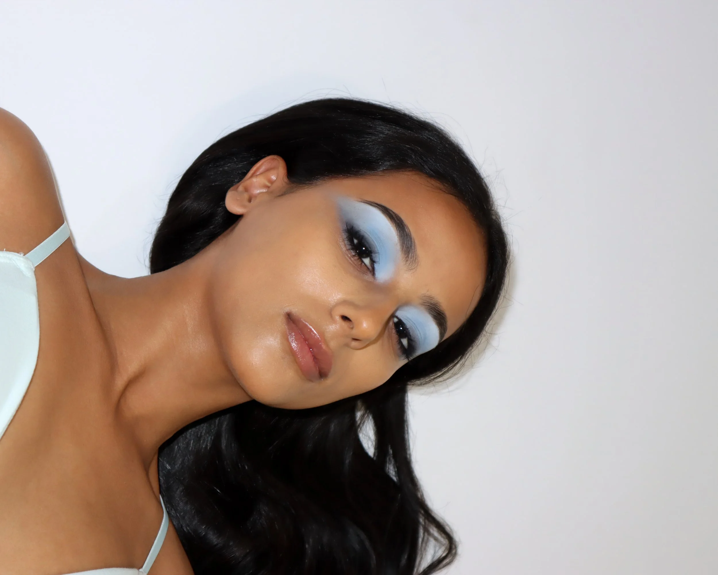 A woman with short black hair and makeup featuring blue eyeshadow, looking sideways, against a plain white background.