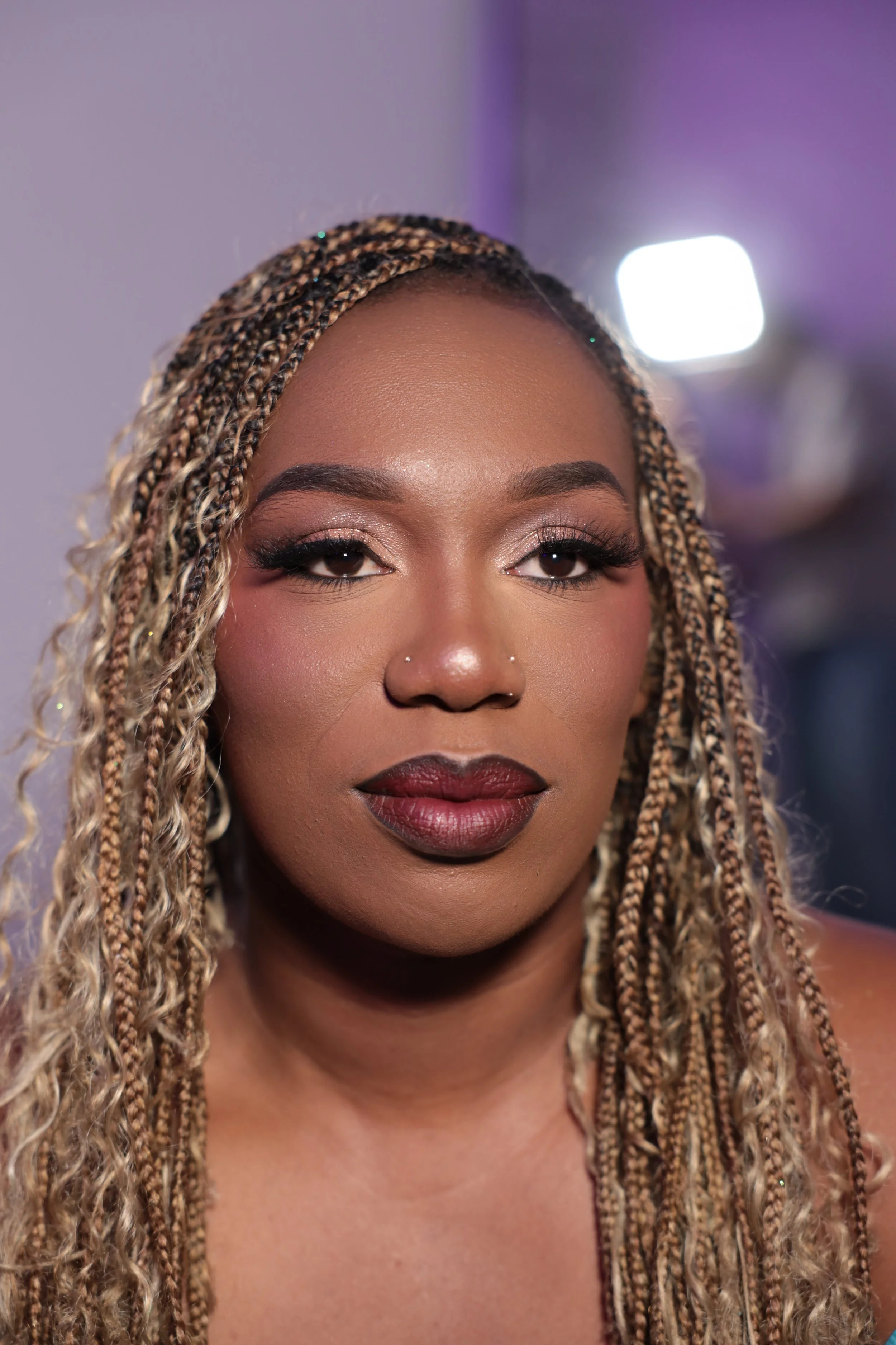 Close-up of a woman with braided hair, makeup, and a nose piercing, looking directly at the camera.