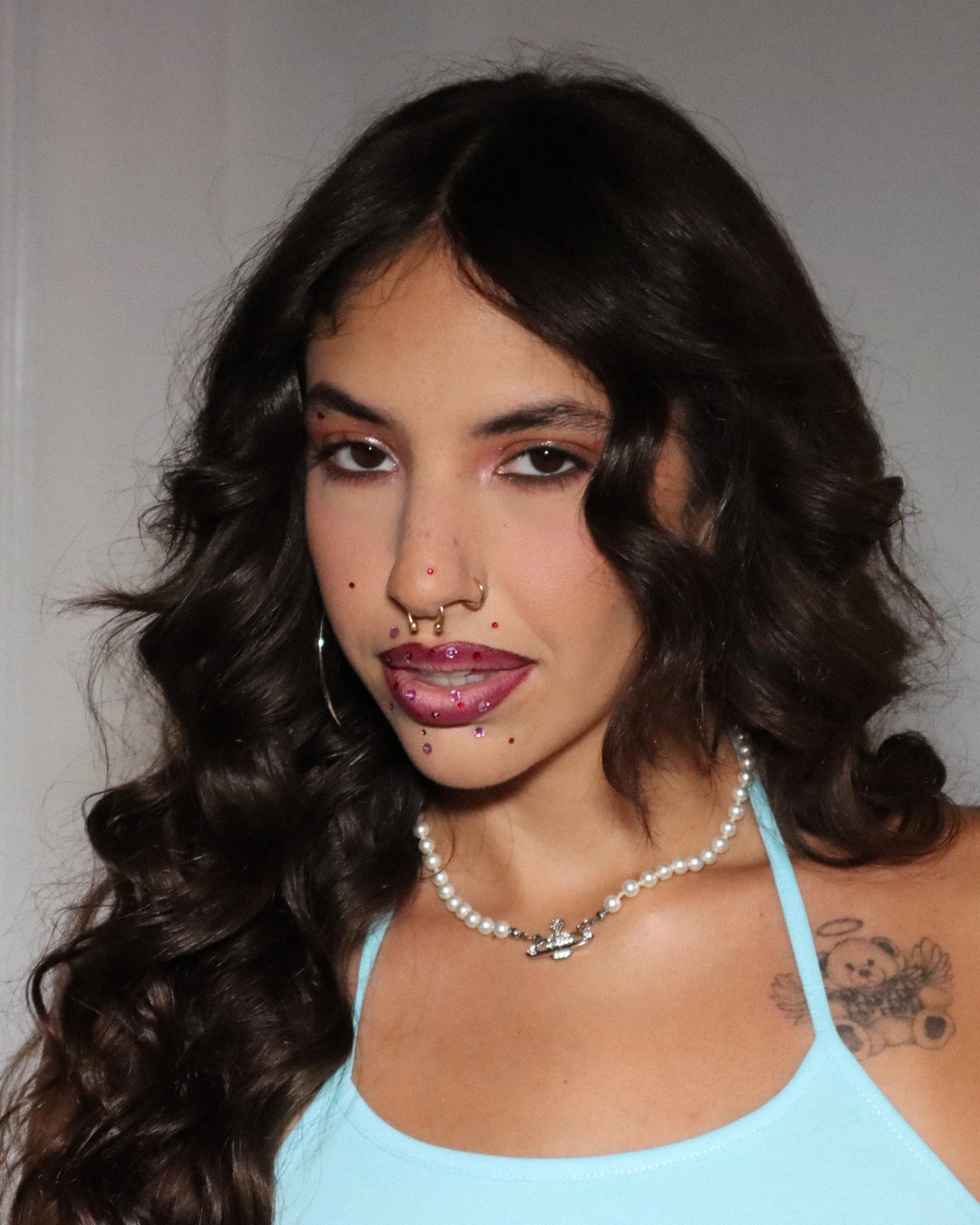Close-up of a woman with dark, curly hair, wearing a light blue top, pearl necklace, and girl makeup, with visible tattoos on her chest.