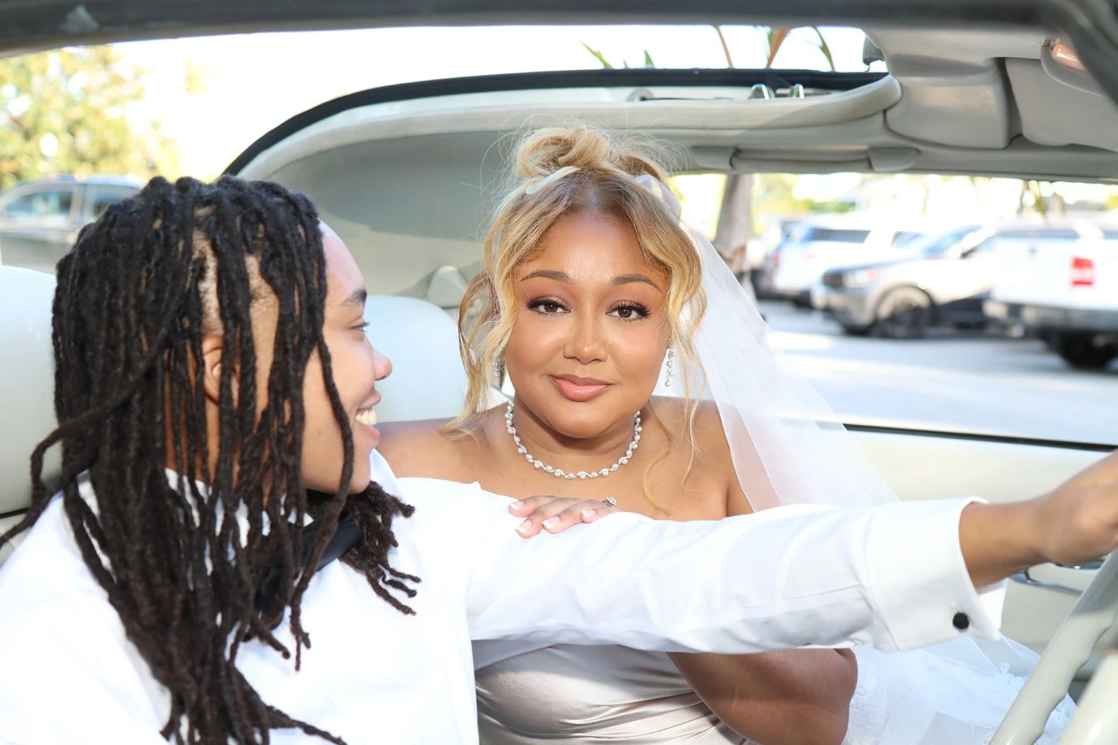 Two women sitting in a car, with one woman dressed as a bride in a wedding gown and veil, and the other woman smiling, taking a selfie together.