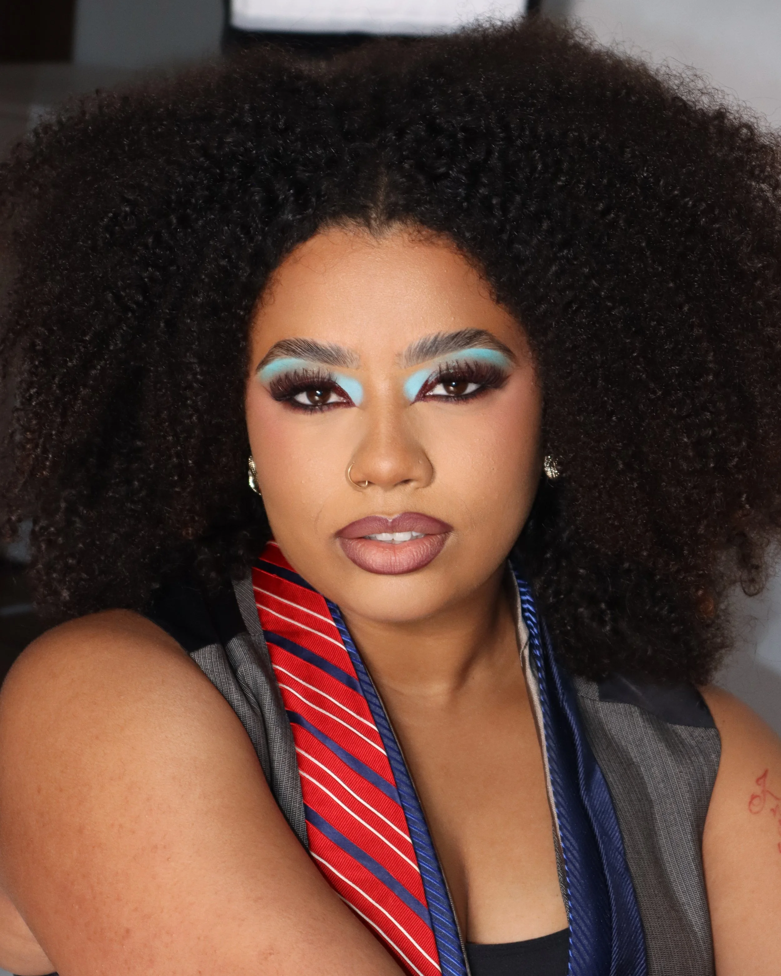 A woman with curly black hair and medium skin tone wearing eye makeup with blue eyeshadow and long eyelashes, a nose ring, and earrings. She is dressed in a professional outfit with a gray vest and a blue striped tie. The image is a close-up of her f