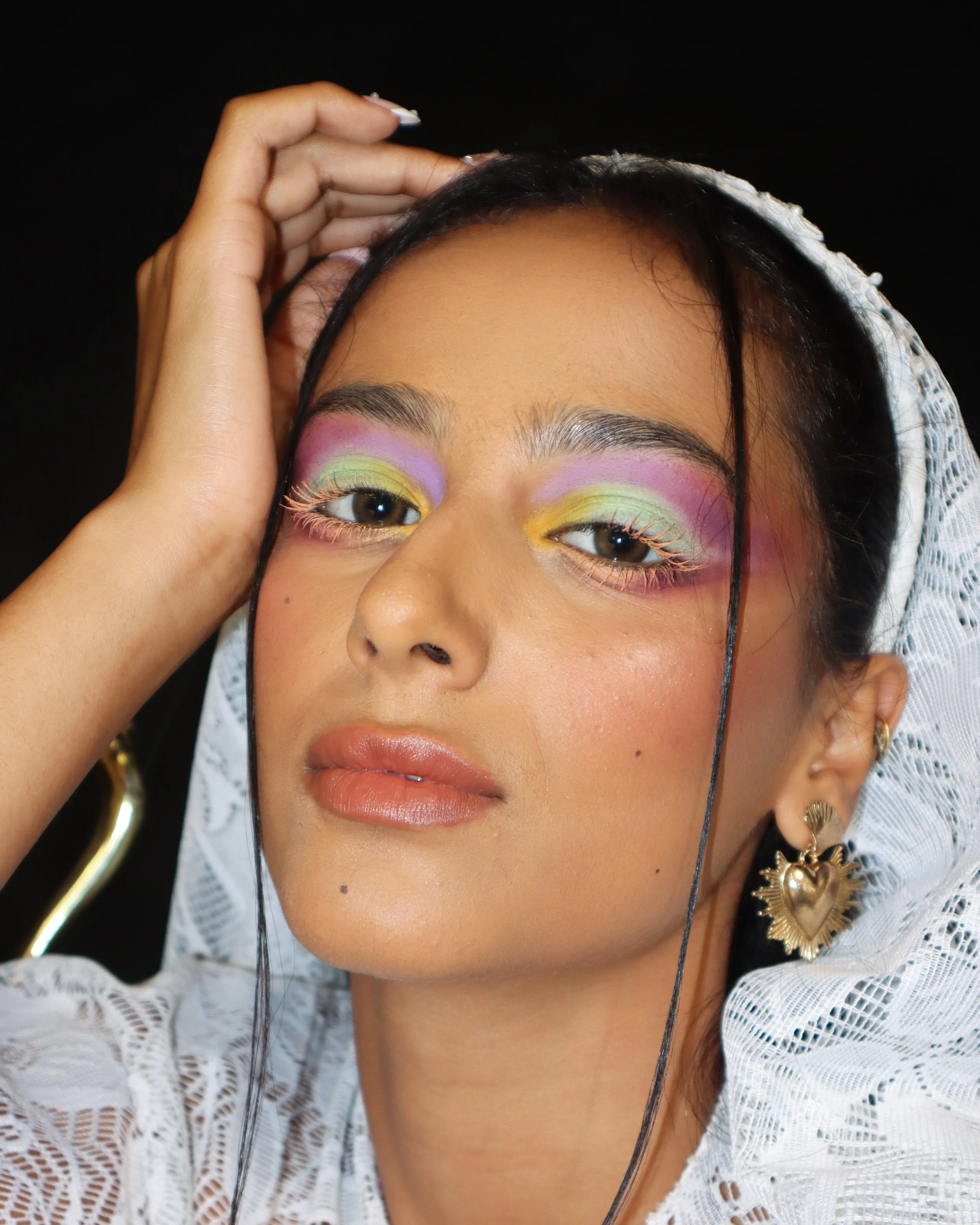 Close-up of a woman's face with colorful pastel eye makeup and gold earrings, wearing a white lace garment and holding her hair back.