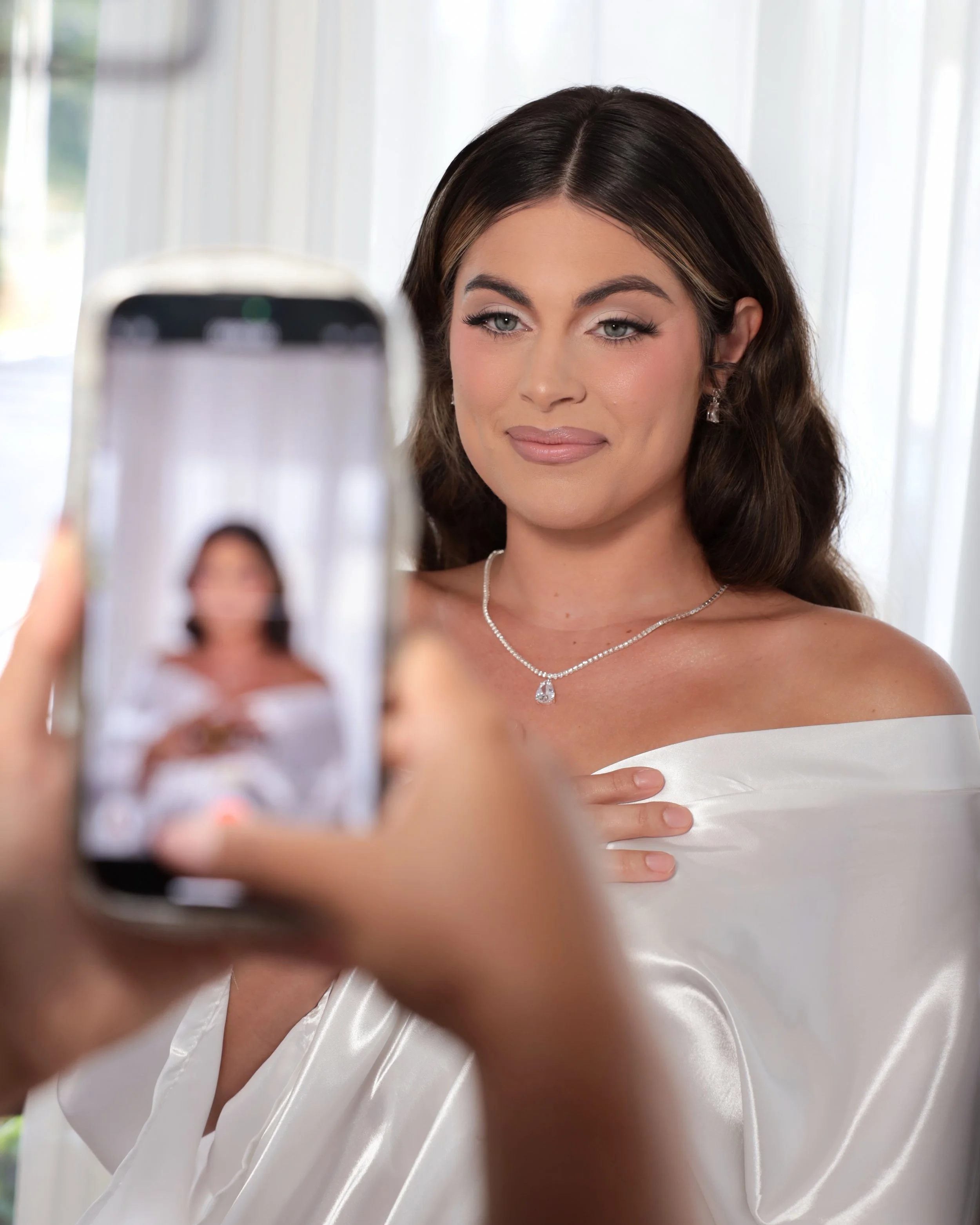 Close-up of a woman with dark brown hair taking a selfie with a smartphone, wearing earrings, a necklace with a teardrop pendant, and an off-shoulder white dress, in a bright indoor setting with curtains in the background.