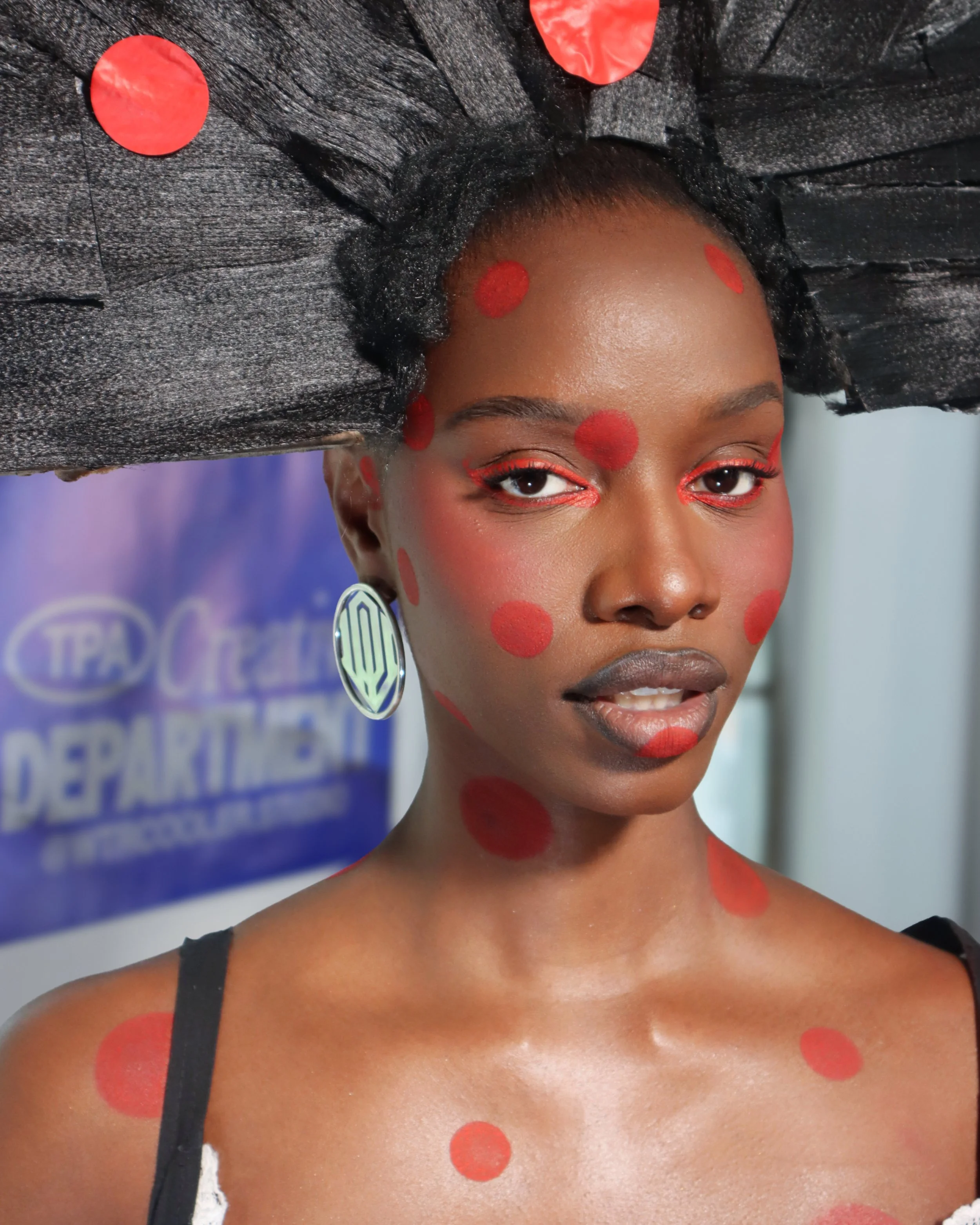 Close-up photo of a woman with dark skin wearing a black dress and large circular earrings. Her face and upper body are painted with red polka dots, and she has red eye makeup. The background shows a partial view of a blue poster with white text.