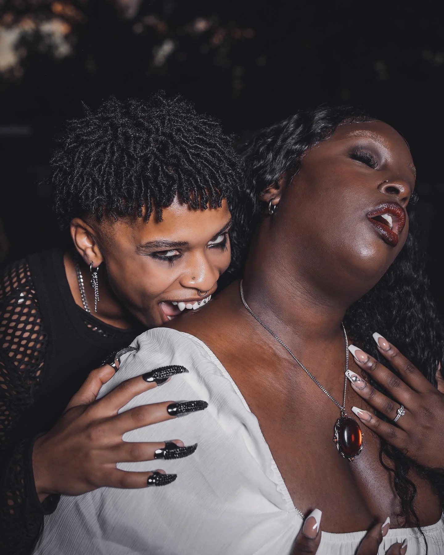 Two women with dark skin tones in an intimate pose, one with short curly hair and the other with long, wavy hair, both accessorized with earrings and necklaces, wearing makeup with dark lipstick, against a dark background.