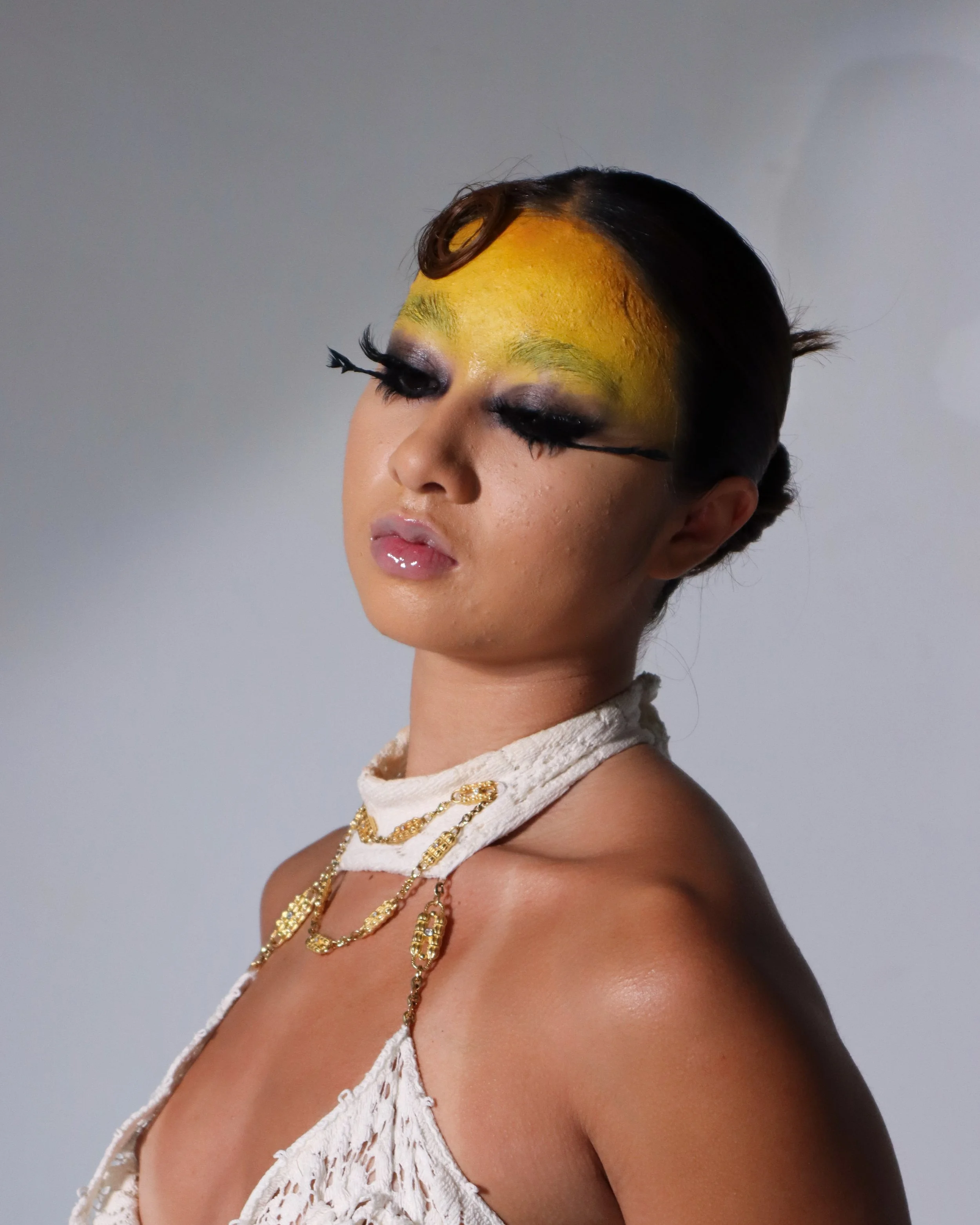 A woman with makeup and jewelry, wearing a white lace garment, poses against a plain background with her eyes closed.