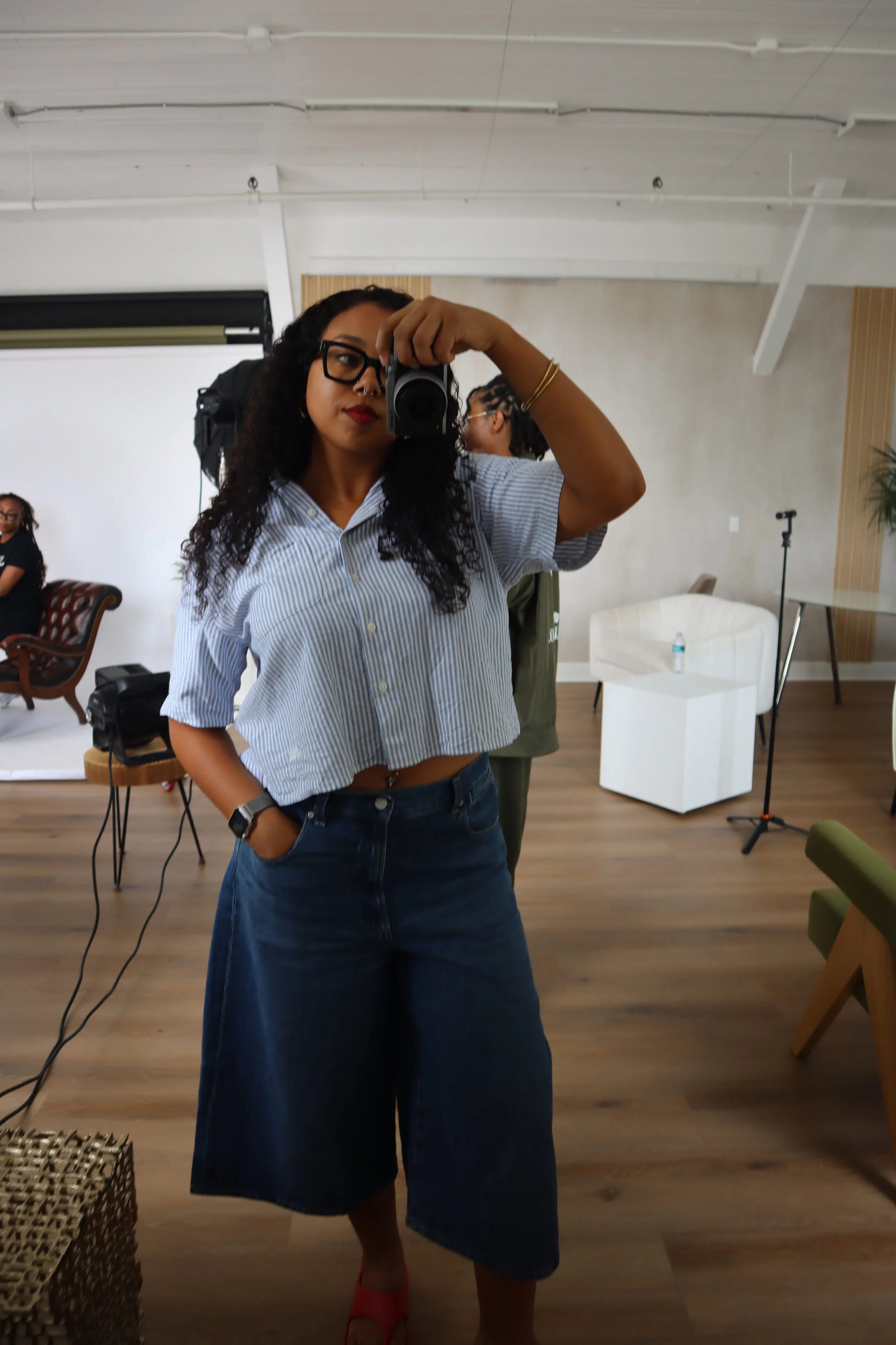 A woman with dark curly hair, glasses, and red lipstick takes a mirror selfie with a camera. She is wearing a light blue striped shirt, dark wide-leg jeans, and red shoes. Makeup artist at a photography studio in Tampa Florida.