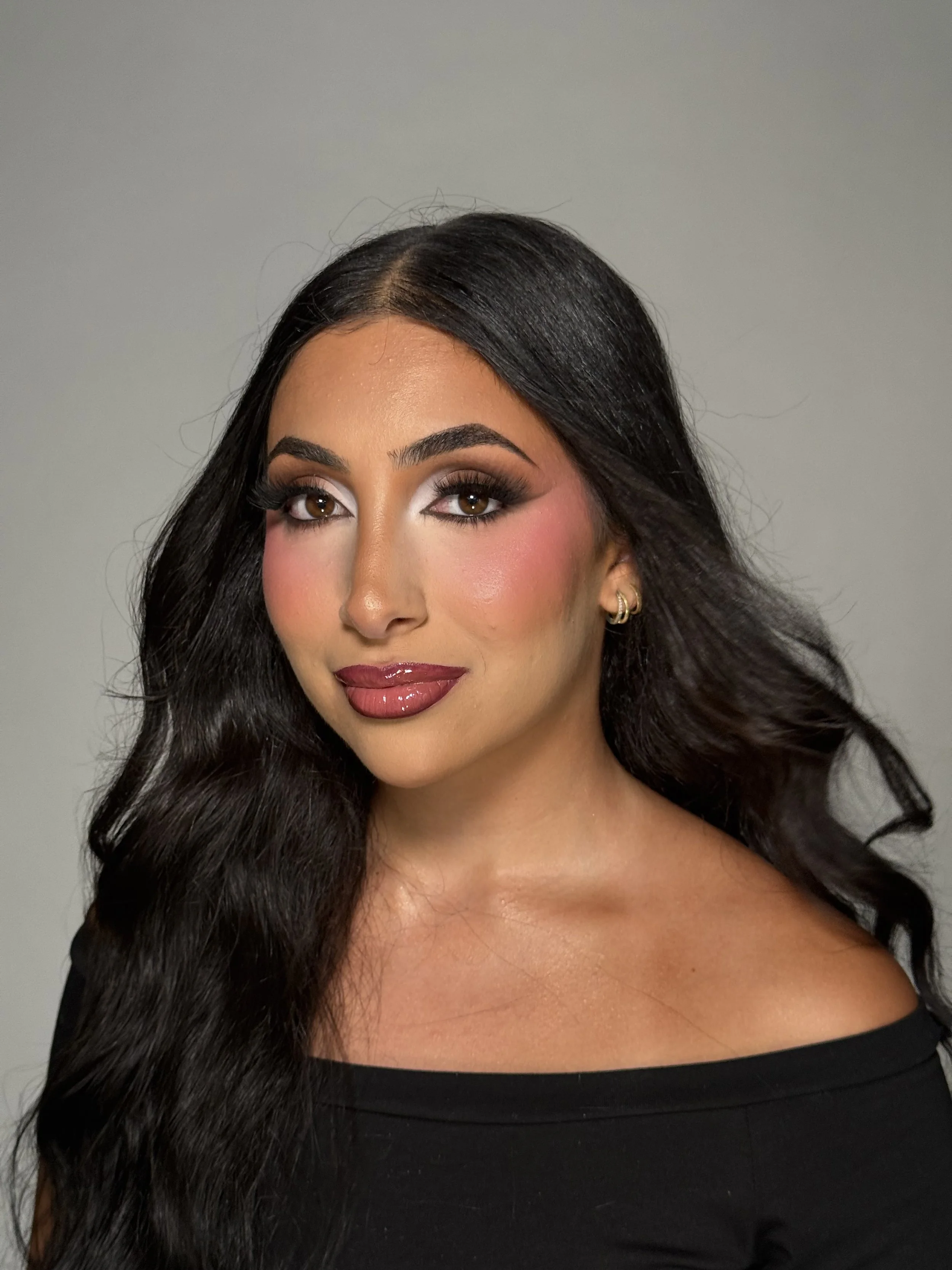 Close-up of a woman with long black hair, makeup including eyeshadow, eyeliner, mascara, blush, and lipstick, wearing a black top with a wide neckline, against a plain gray background.