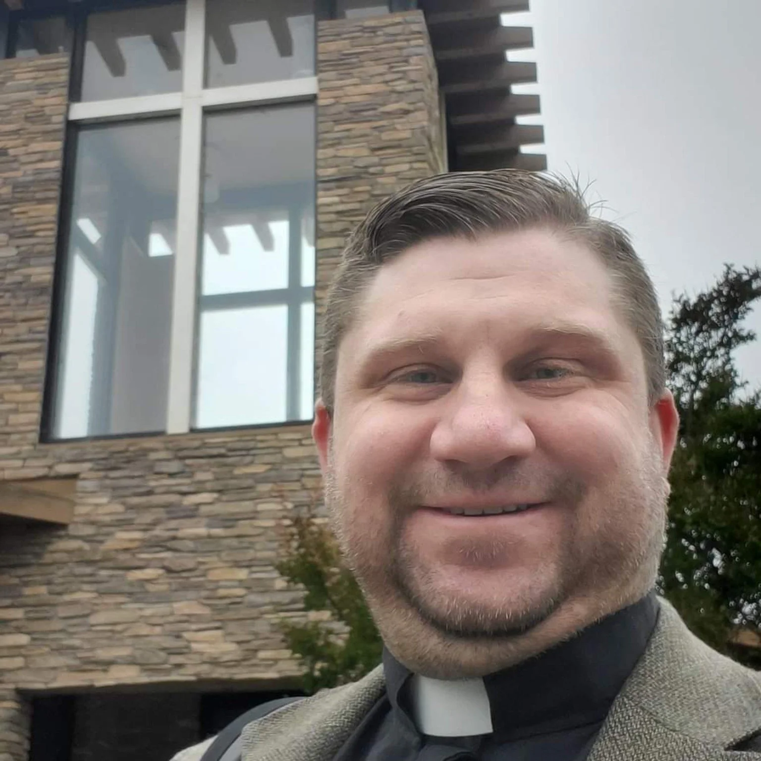 Brian Brozovic wearing a clerical collar outside a modern church building.