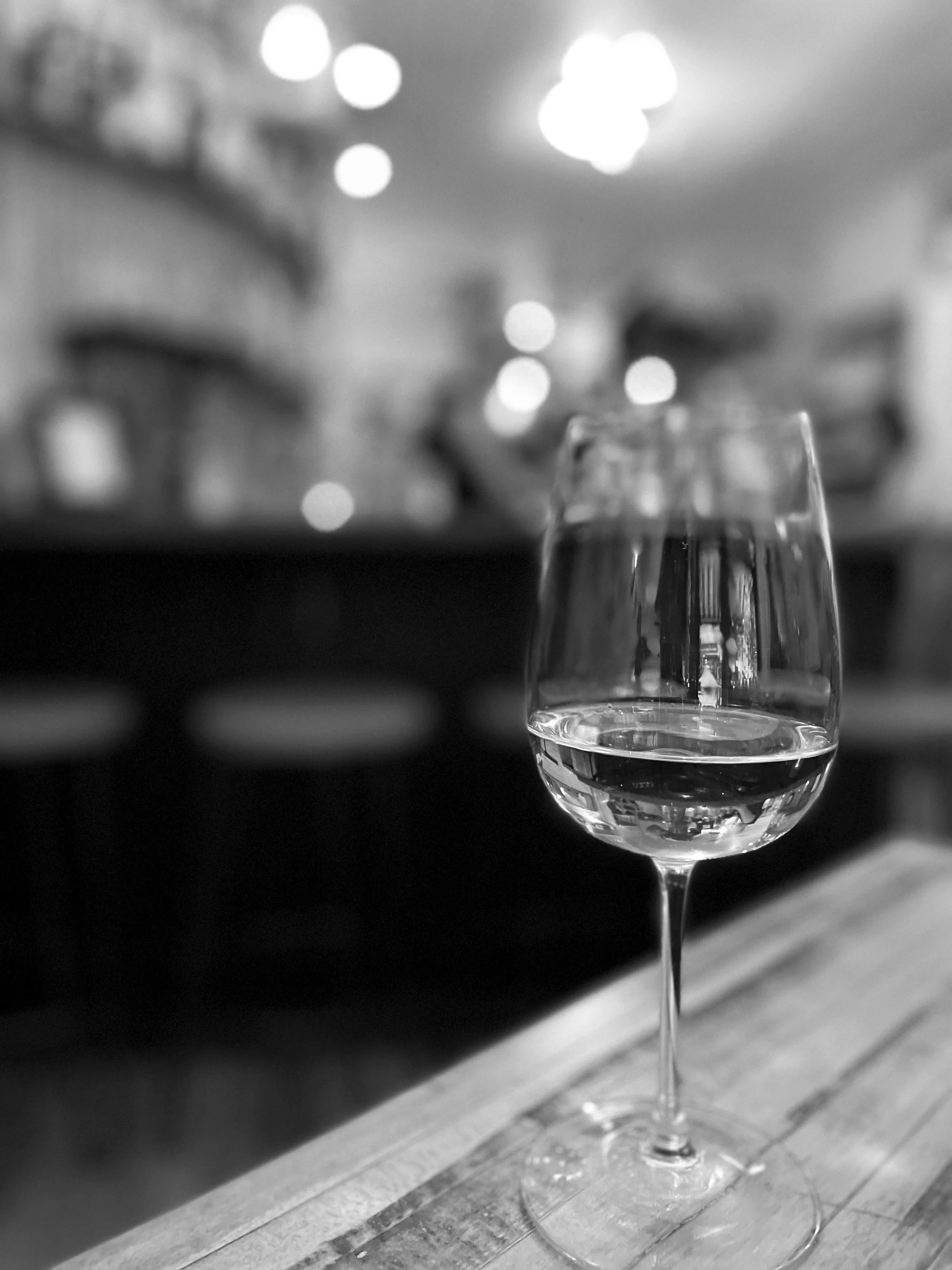 Black-and-white photograph of a glass of white wine on a restaurant bar with softly blurred lights in the background