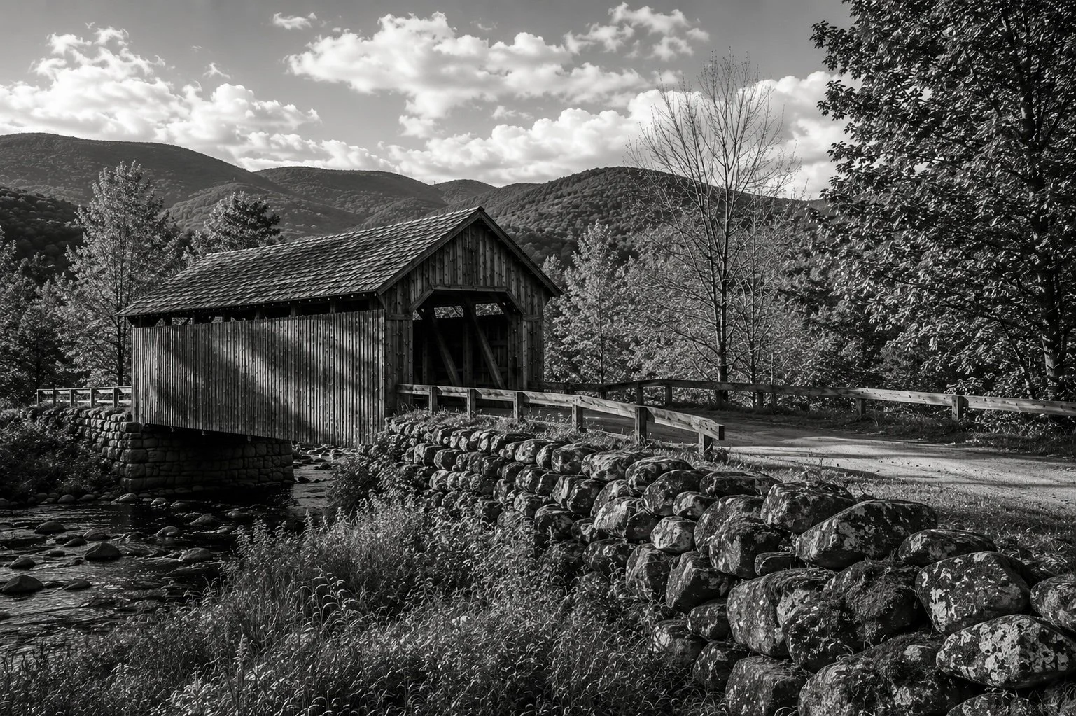 Black-and-white photograph of a covered bridge beside a stone wall in a mountainous New England landscape