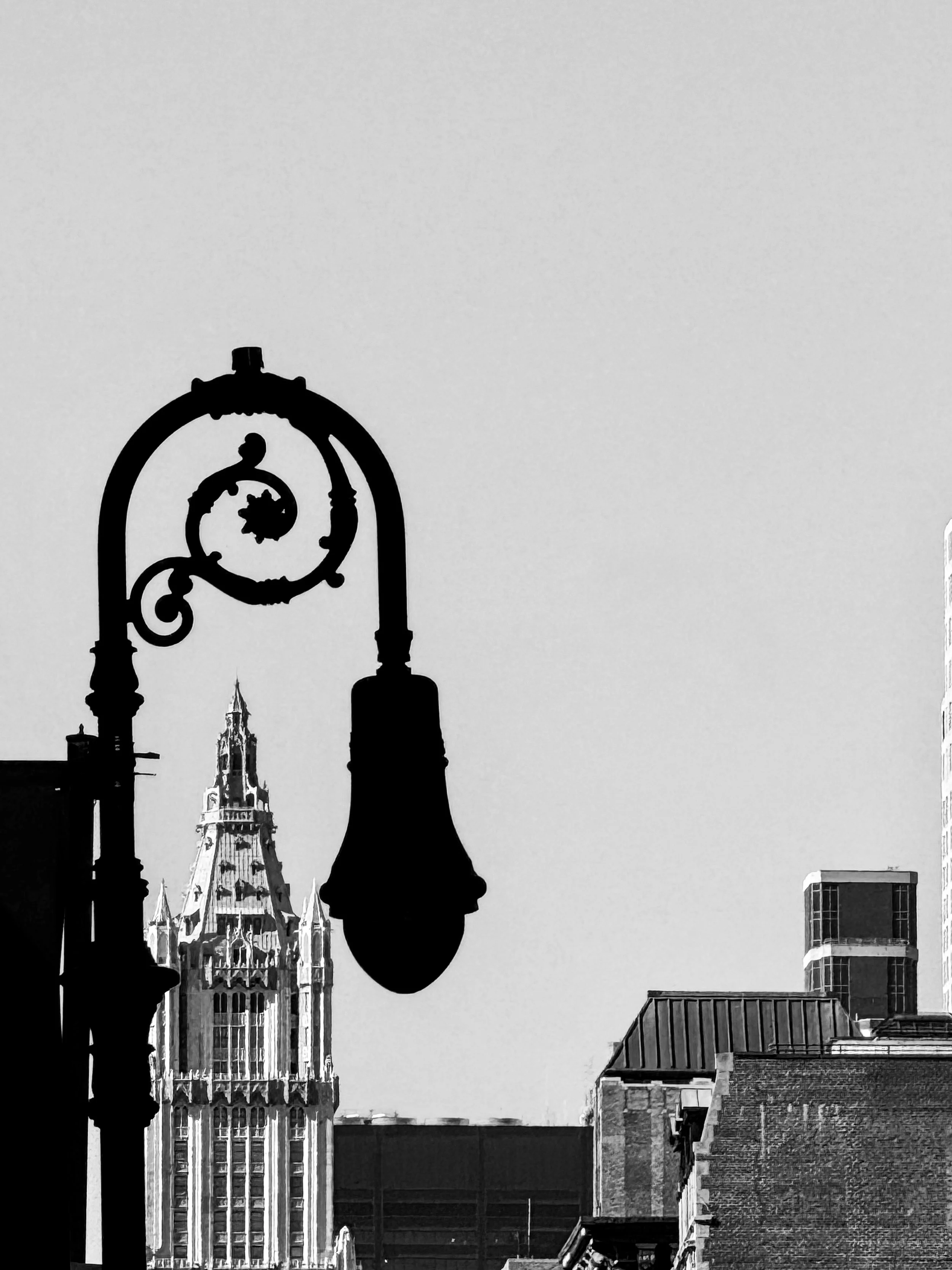 Black-and-white photograph of the Woolworth Building framed by an ornate street lamp and Manhattan rooftops
