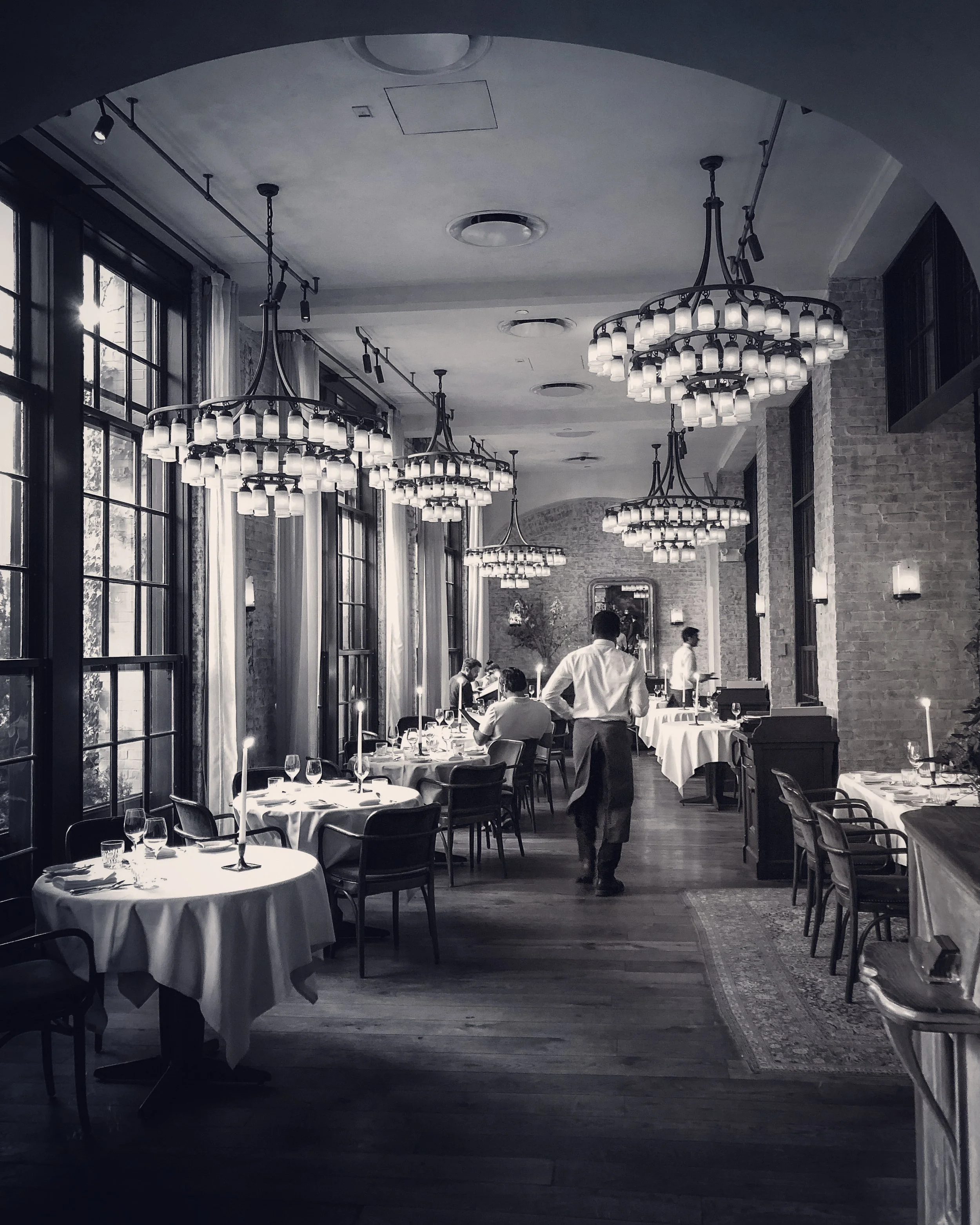 Black-and-white photograph of the dining room at Le Coucou in Manhattan, with chandeliers and set tables