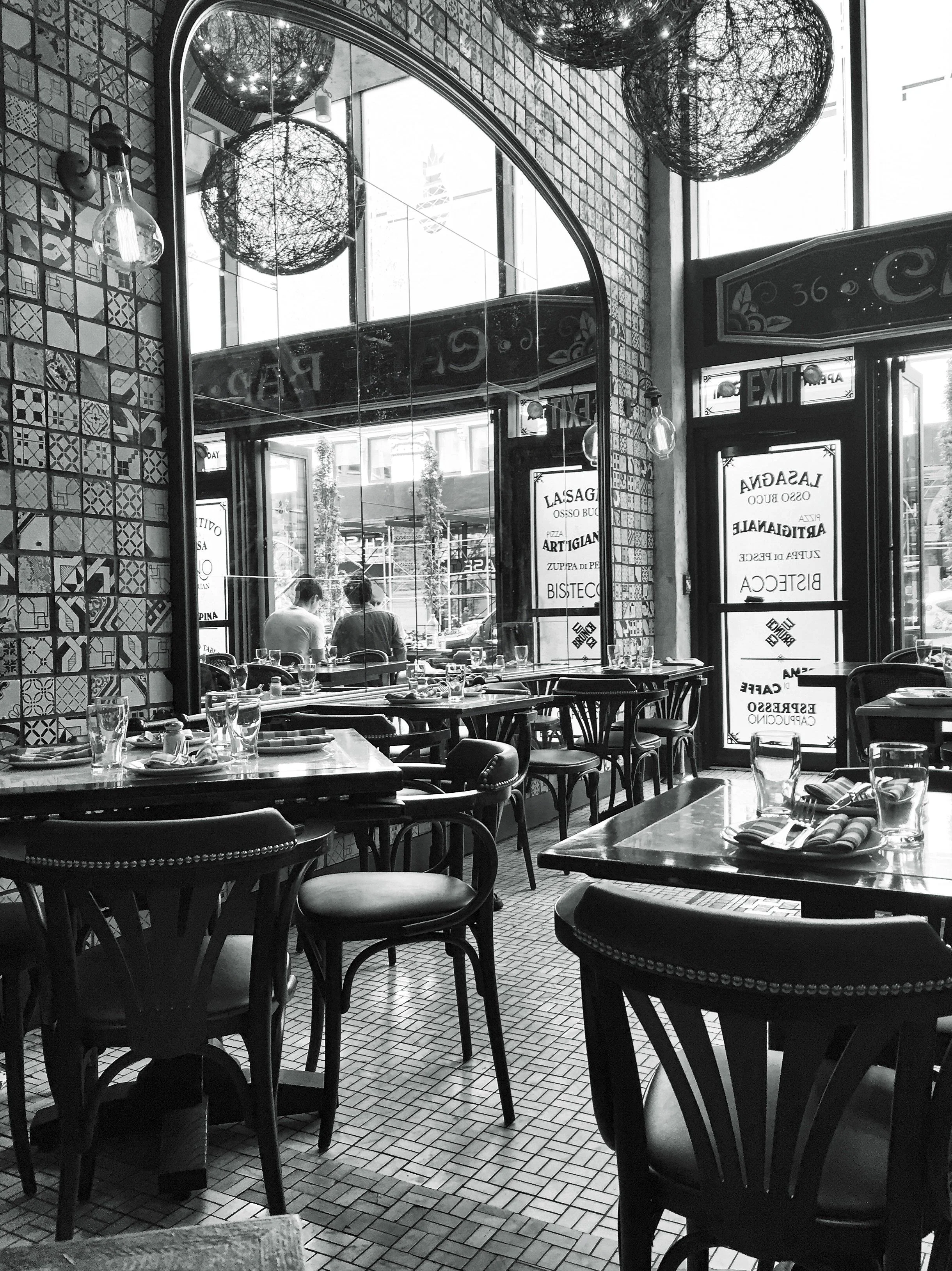 Black-and-white photograph of a French restaurant dining room with arched windows, hanging globe lights, and set tables