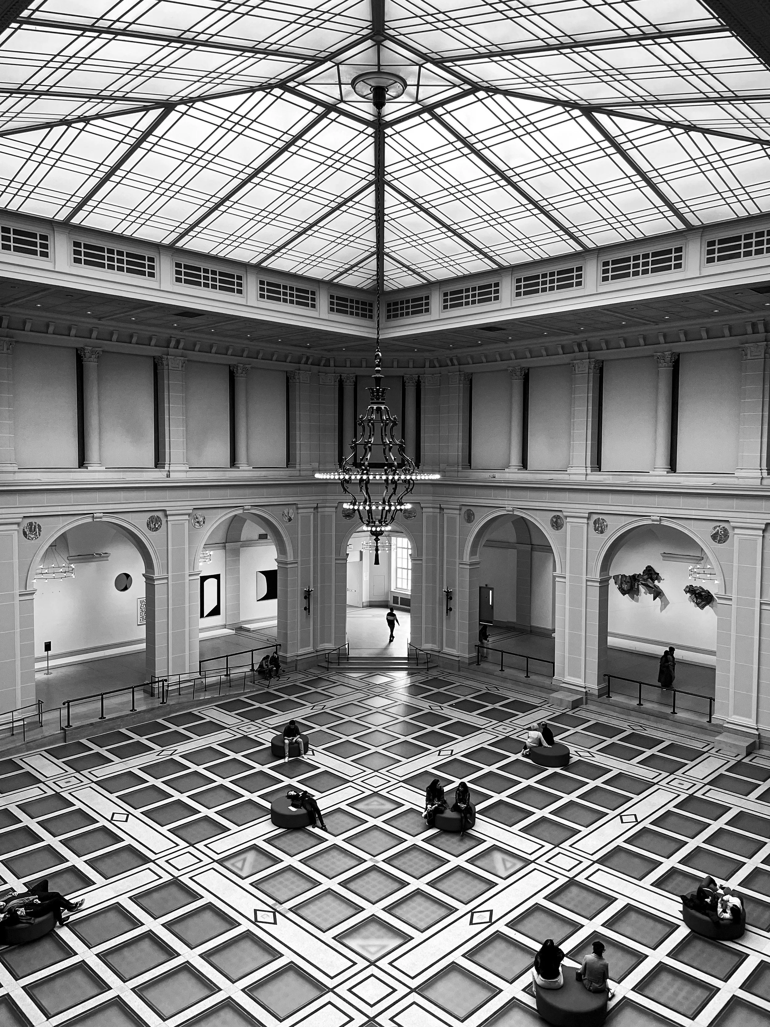 Black-and-white photograph of the Brooklyn Museum's grand interior court, with a geometric patterned floor, arched doorways, chandelier, and glass ceiling