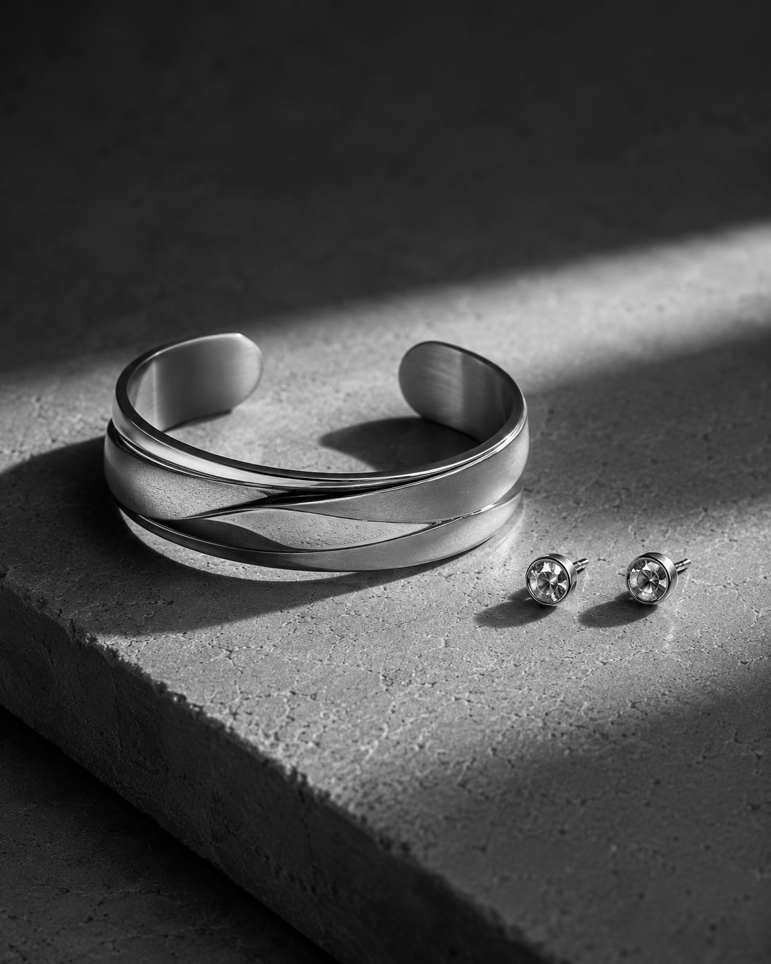 Black-and-white photograph of a sculptural metal cuff bracelet and a pair of stud earrings illuminated by a beam of light