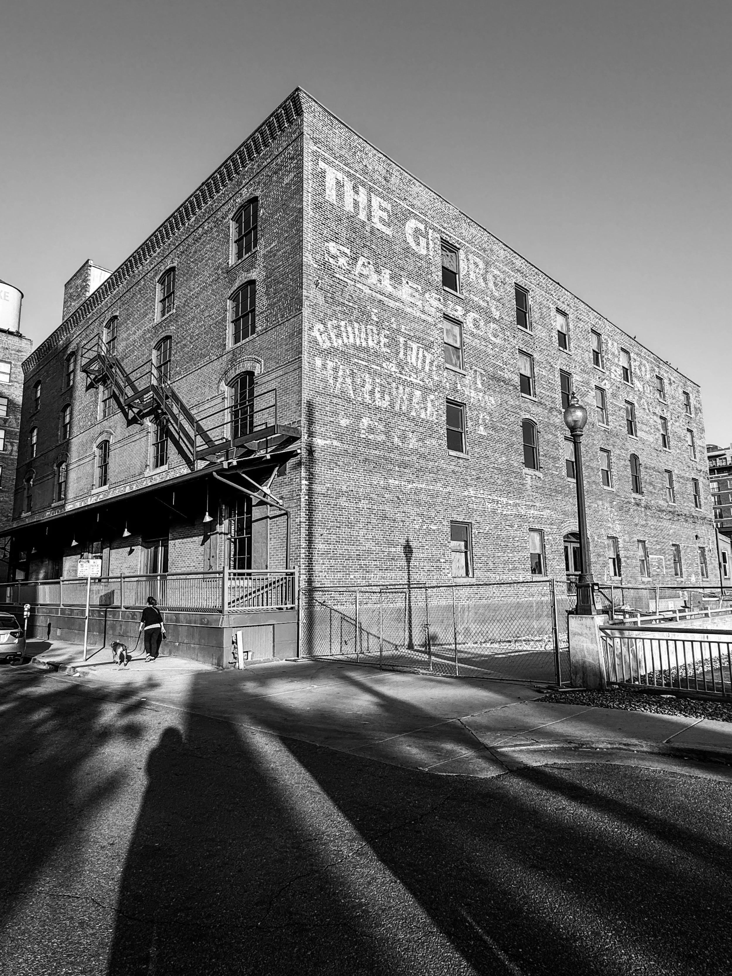 Black-and-white photograph of a large brick industrial building with faded painted signage and long shadows across the street in Denver Colorado