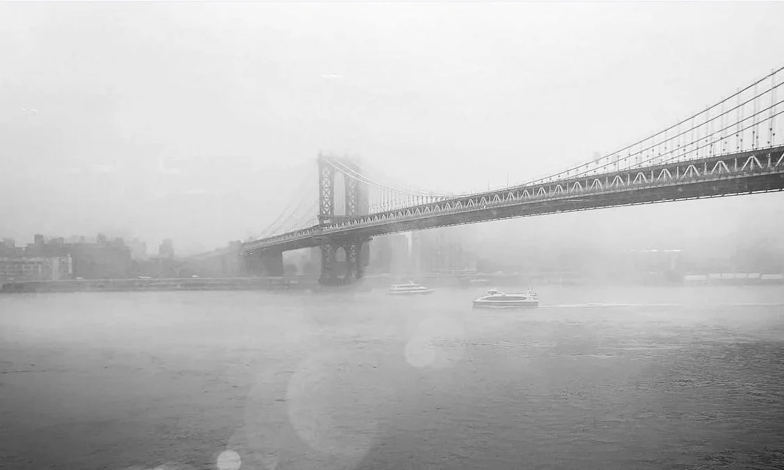 Black-and-white photograph of the Manhattan Bridge emerging through mist, viewed from DUMBO, Brooklyn