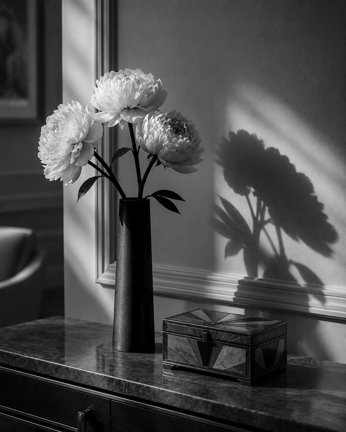 Black-and-white still life of flowers in a tall vase beside art deco box casting shadows on the wall