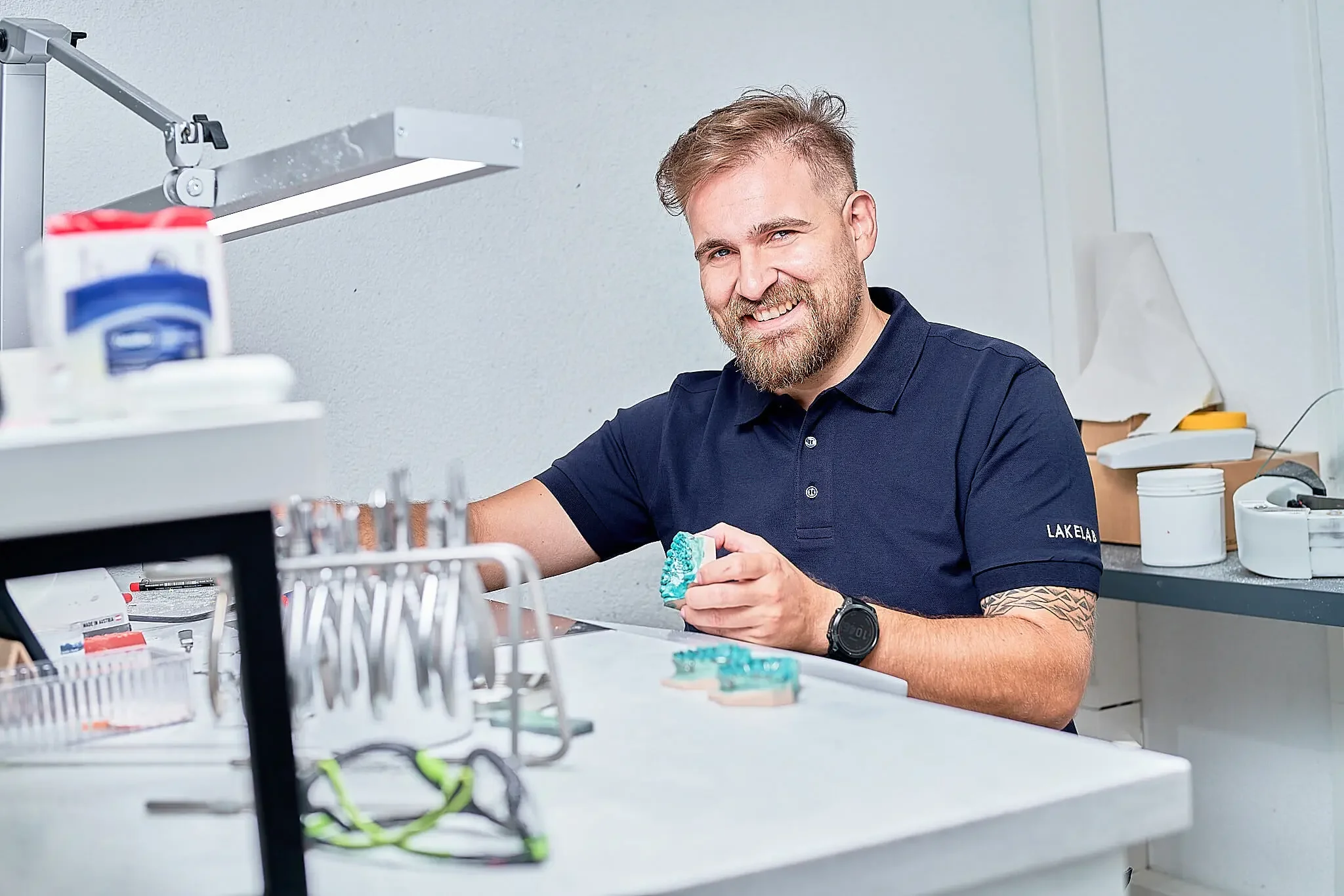Lächelnder Mann mit Bart und Tattoo am Arm sitzt an einem Laborarbeitsplatz, hält ein Objekt in der Hand und trägt ein schwarzes Polo-Shirt mit der Aufschrift 'LAK ELAB'.