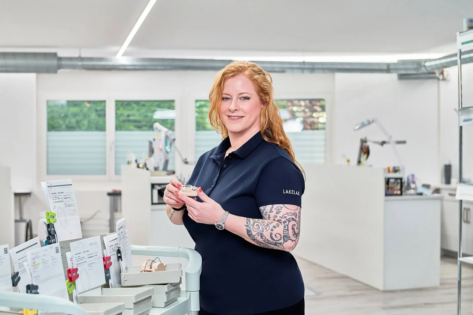 Eine Frau mit roten, lockigen Haaren und Tattoos auf den Armen, trägt eine schwarze Kleidung mit der Aufschrift 'LAKELAB' und hält eine Brille in den Händen. Sie steht in einem hellen, modernen Labor oder Arbeitsraum mit Werkzeugen und medizinischen Artikeln auf einem Tisch.