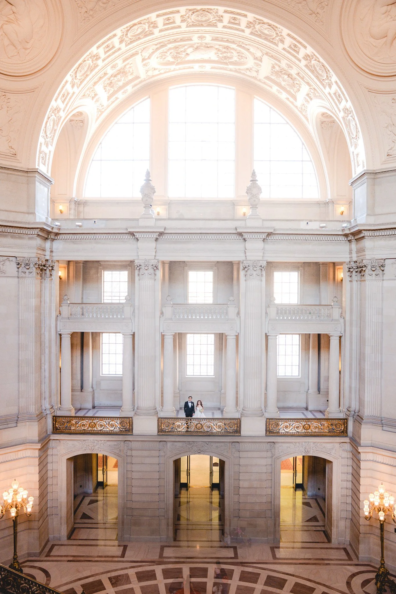 A bride and groom standing together in a grand, elegant interior with large arched windows, ornate details, and columns, inside of San Francisco City Hall. 