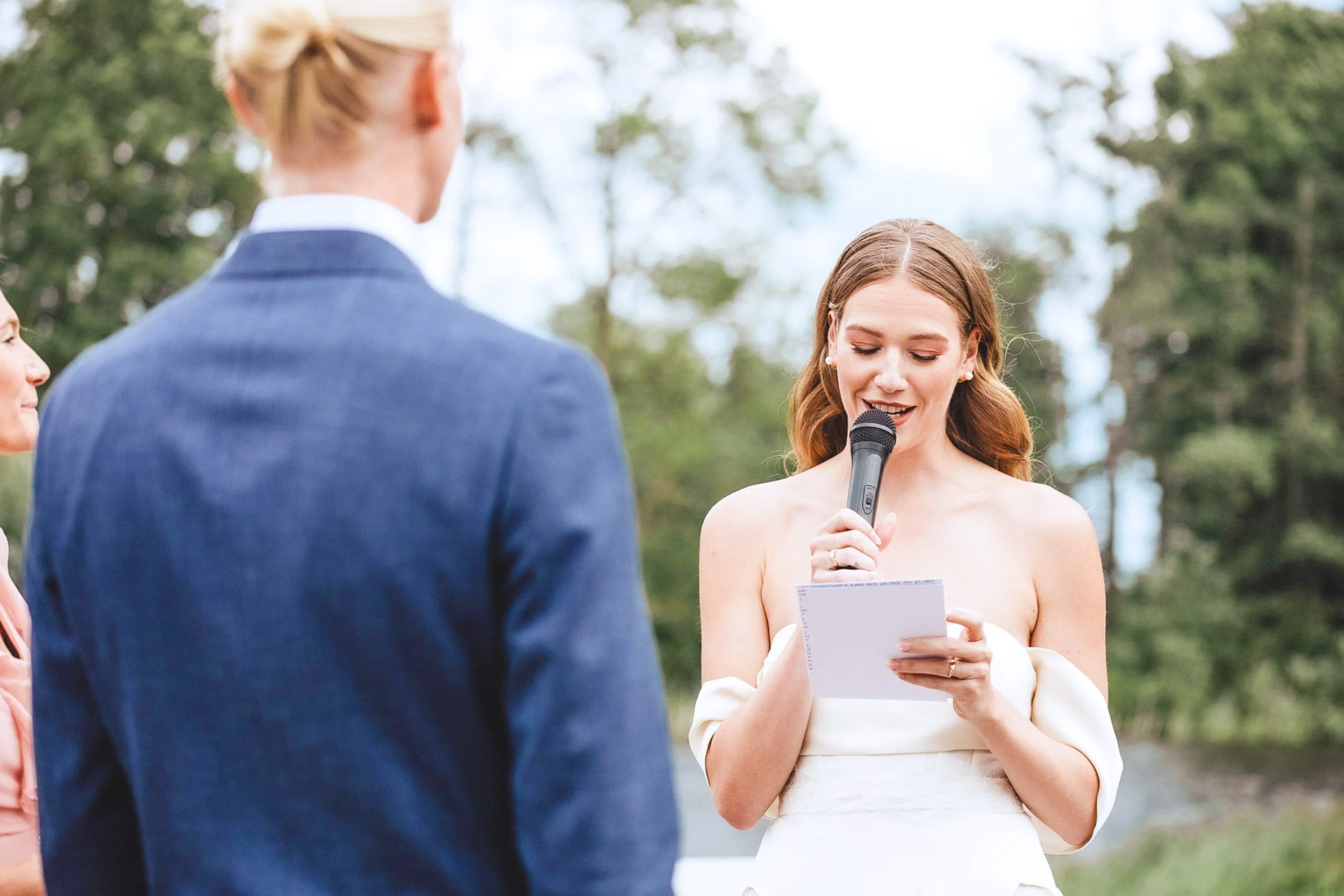 A woman in a wedding dress holding a microphone and reading from a piece of paper during an outdoor wedding ceremony, with a man in a suit standing in front of her. South Bay.