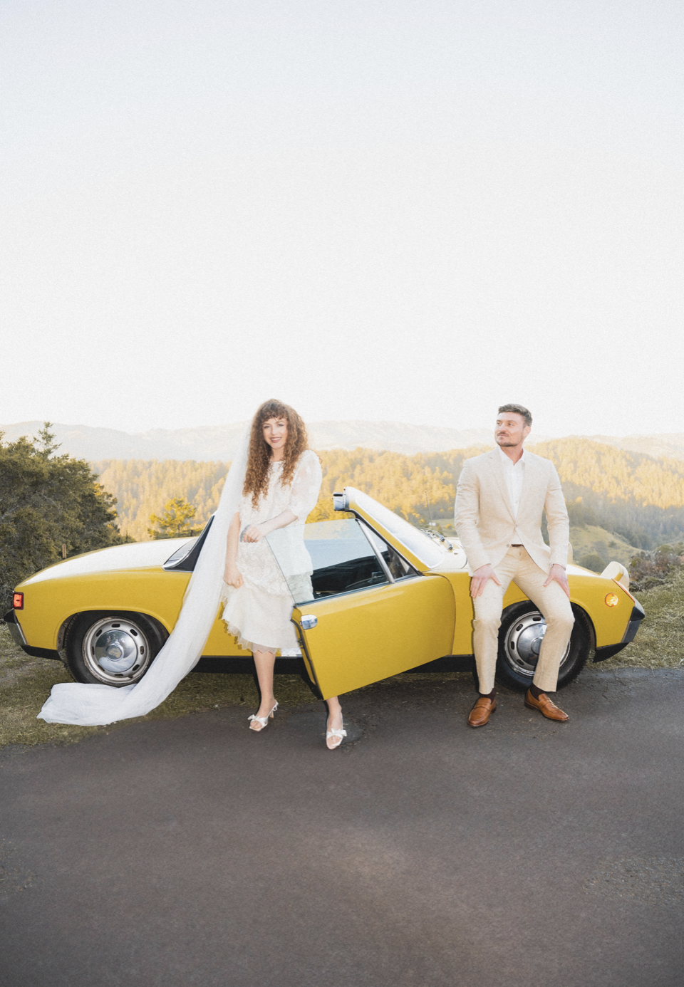 A bride in a white dress and veil standing next to a yellow vintage car, with a groom in a cream suit sitting on the car's edge, outdoors during sunset with a scenic background of Mt Tamalpais hills in Marin County