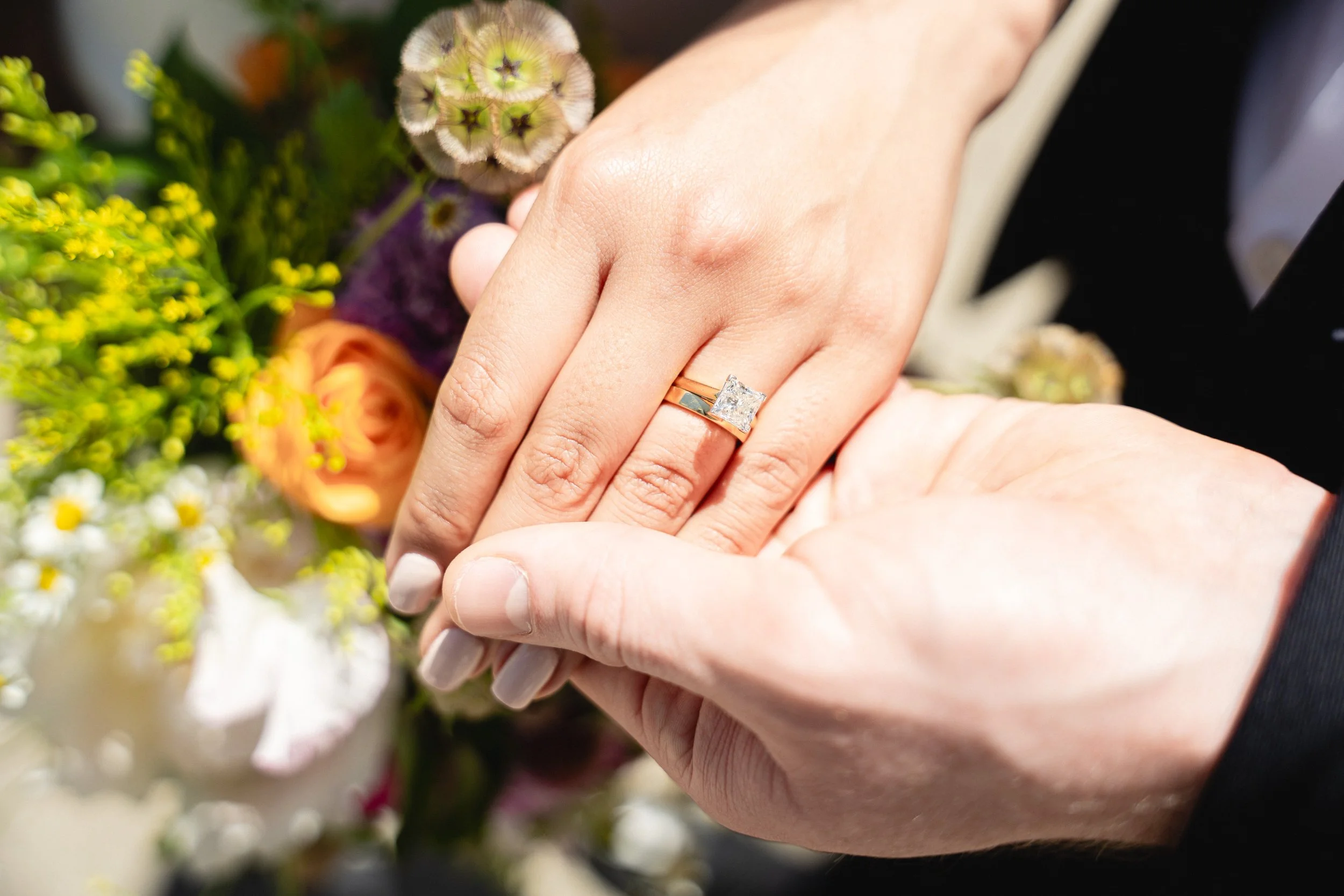 Close-up of two hands holding each other, with one hand wearing a ring with a large diamond. Couple holding hands in front of San Francisco City Hall.