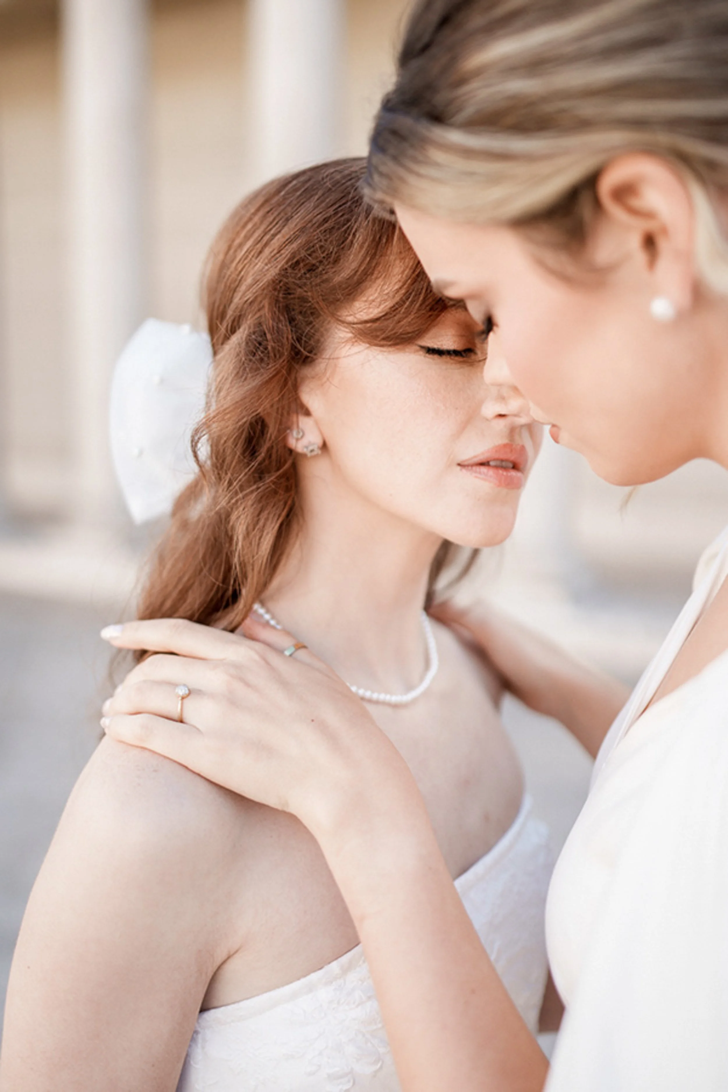 Close-up of a bride and her partner touching foreheads, with the bride in a white dress, wearing pearl jewelry, and her partner in a suit. The moment is tender and intimate. Location is Legion of Honor. LGBTQ+.