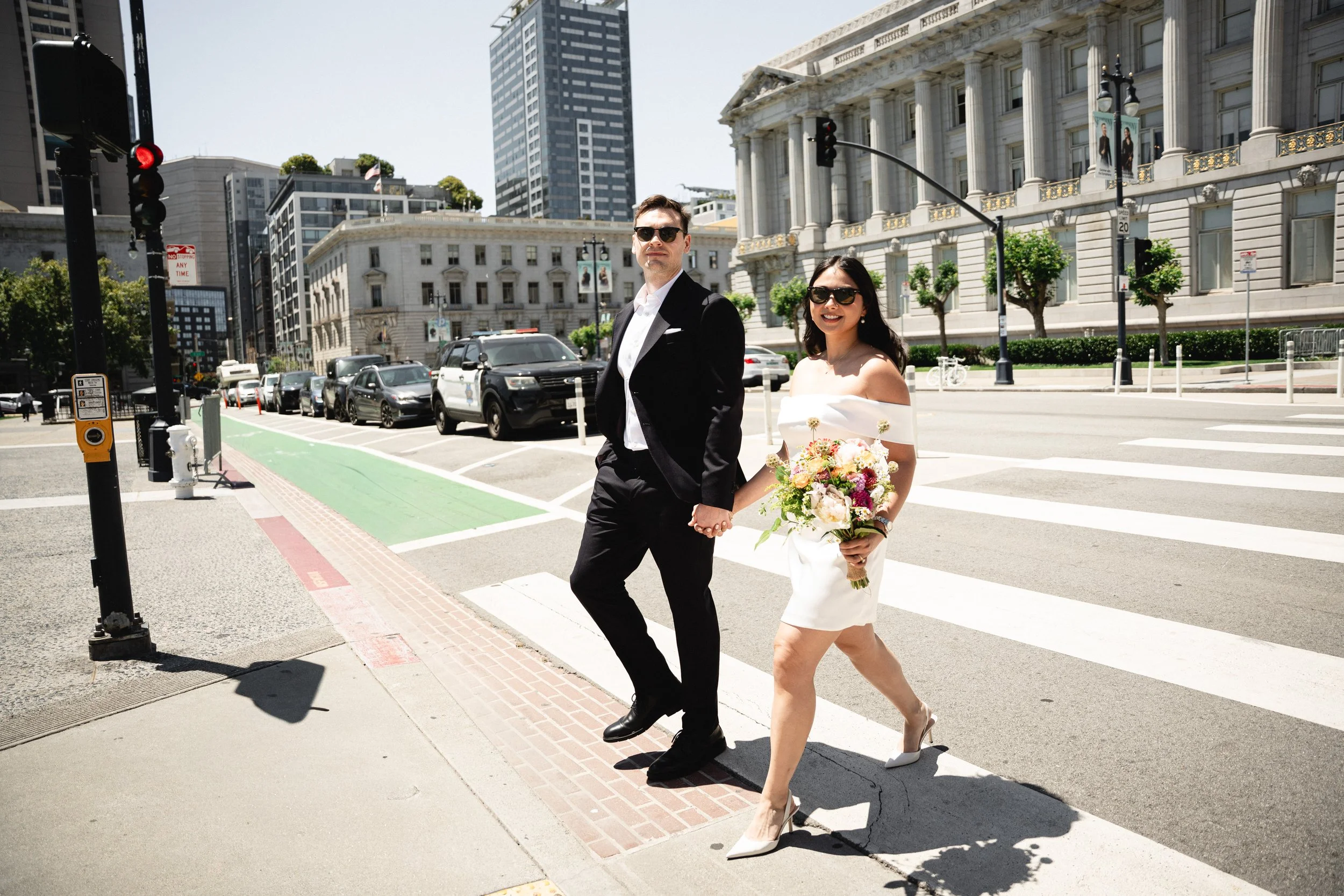 A man in a black tuxedo and a woman in a white dress holding a bouquet crossing a city street on a crosswalk, across the street from SF City Hall 1 Dr Carlton B Goodlett Pl, San Francisco, CA 9410