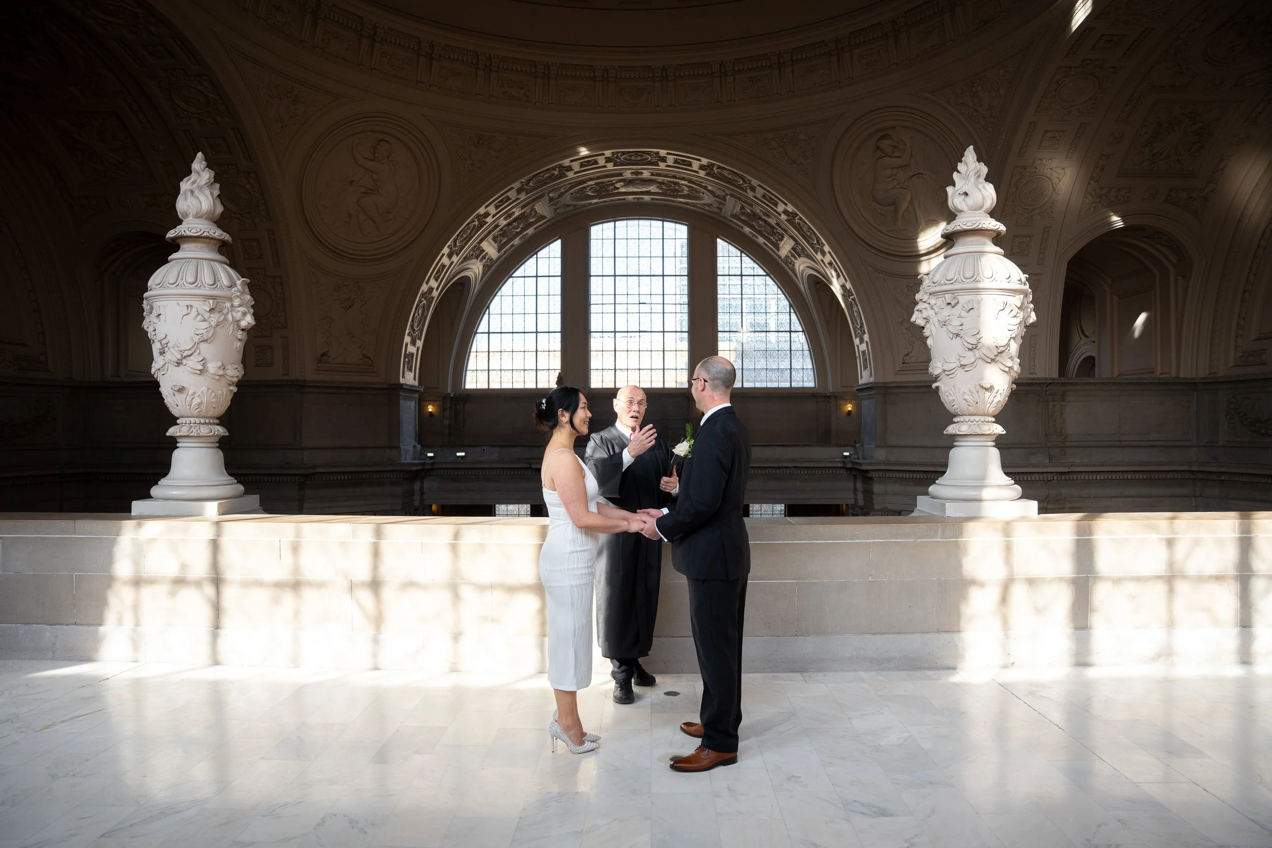 A bride and groom standing face-to-face holding hands during their wedding ceremony inside SF City Hall 1 Dr Carlton B Goodlett Pl, San Francisco, CA 94102. An officiant is speaking to them.