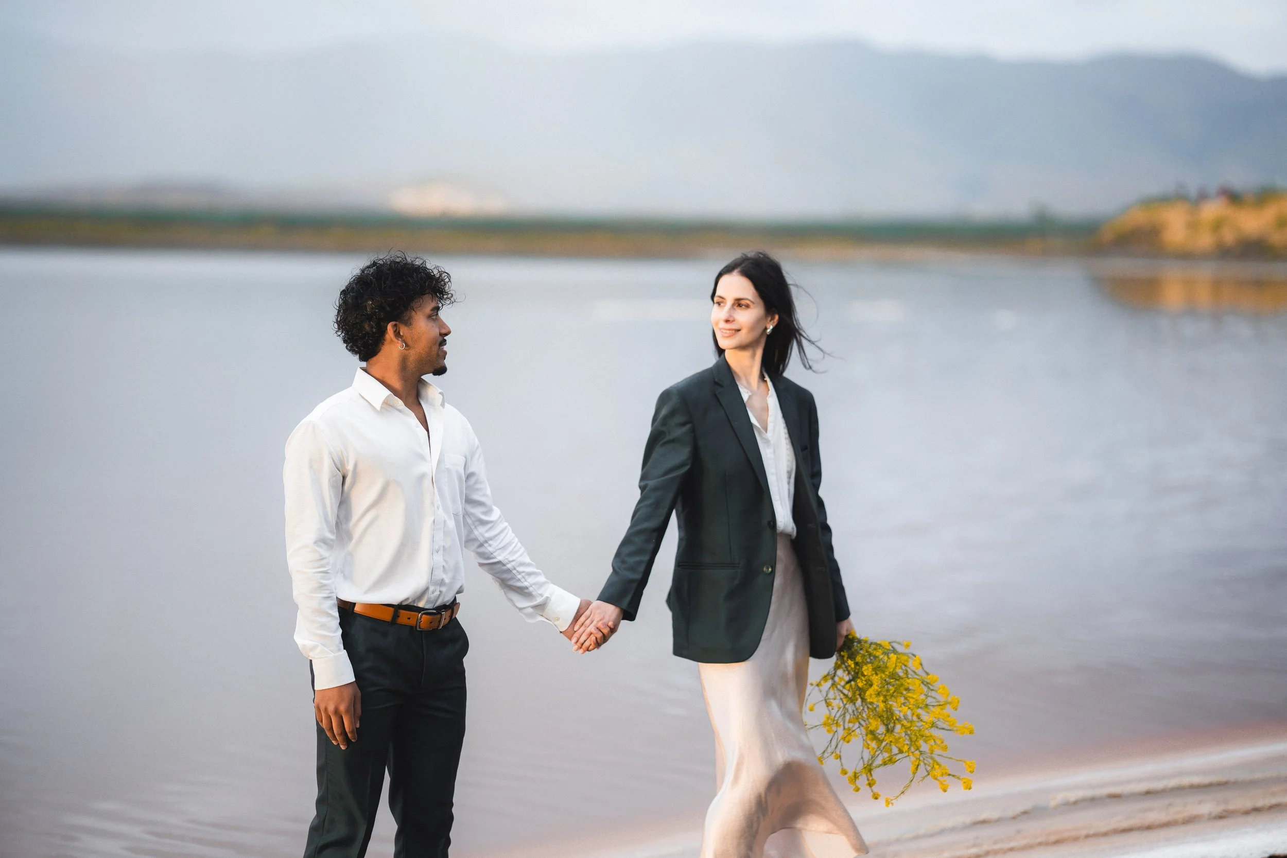 A man and woman holding hands outdoors near a body of water, with the woman holding a bouquet of yellow flowers, and the woman is smiling while looking at the man, near pink lake in Marina Alviso.