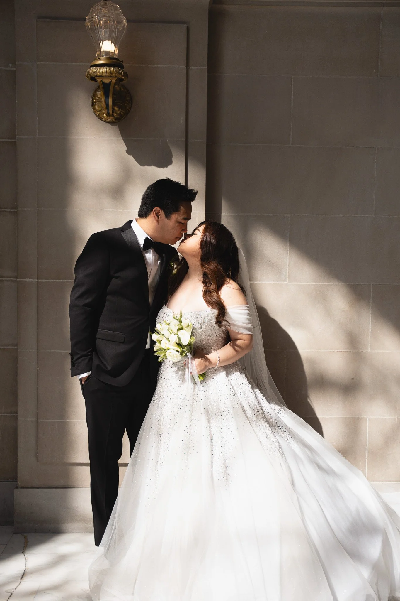 A bride and groom sharing a kiss in front of a grey tiled wall with a vintage wall sconce inside SF City Hall 1 Dr Carlton B Goodlett Pl, San Francisco, CA 94102.