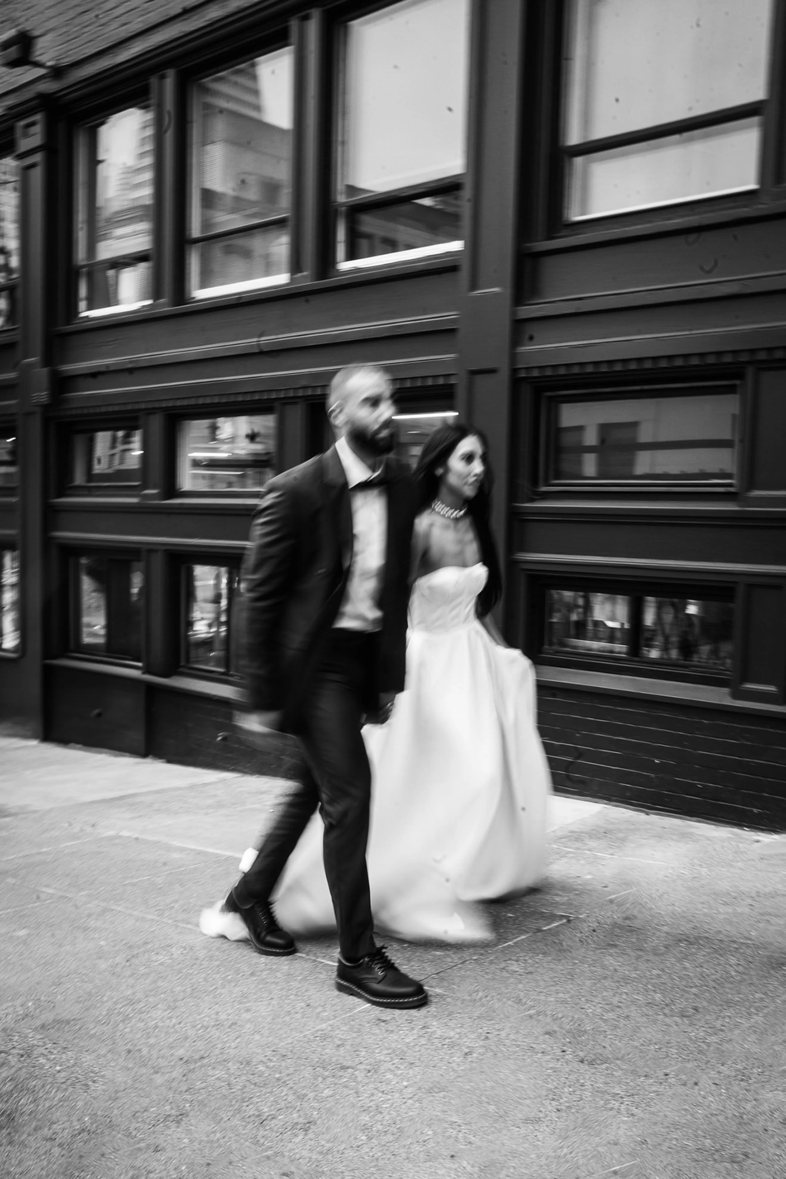 A black and white photo of a man and woman dressed in formal attire walking on a city sidewalk in front of a building with large windows in downtown San Francisco.