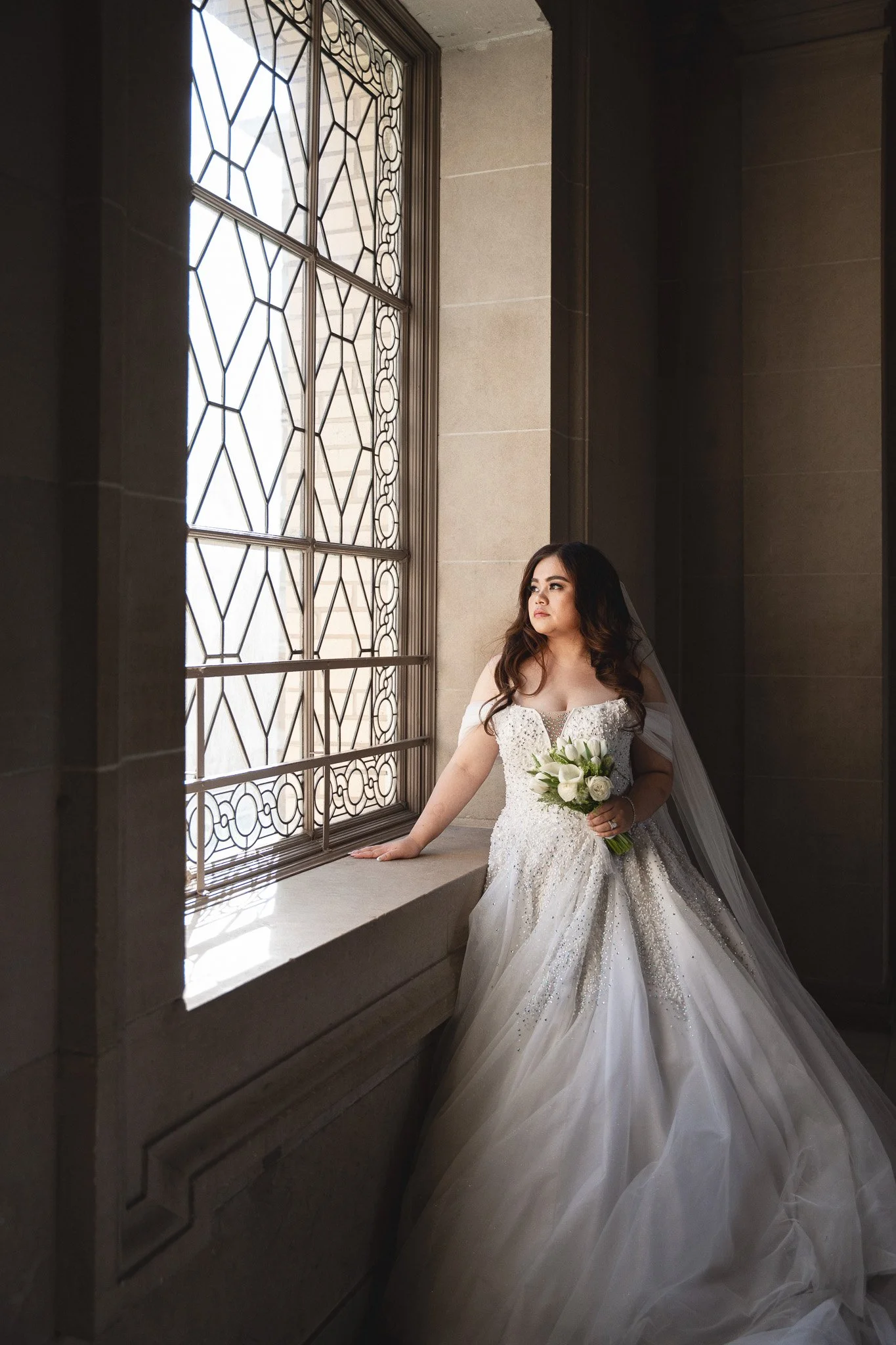 Bride in a white gown gazing out a window near City Hall's ornate ironwork, bouquet in hand.