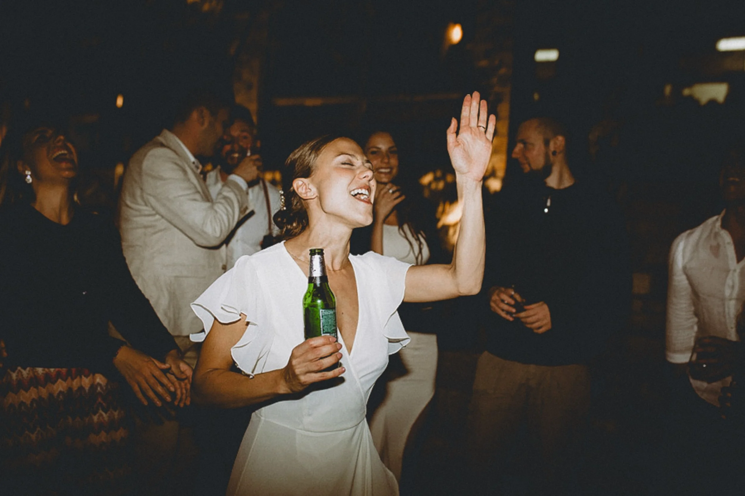 A group of people dancing and laughing at a party, with a woman in the foreground holding a beer bottle, eyes closed, and her right hand raised in celebration. Flash night photography.