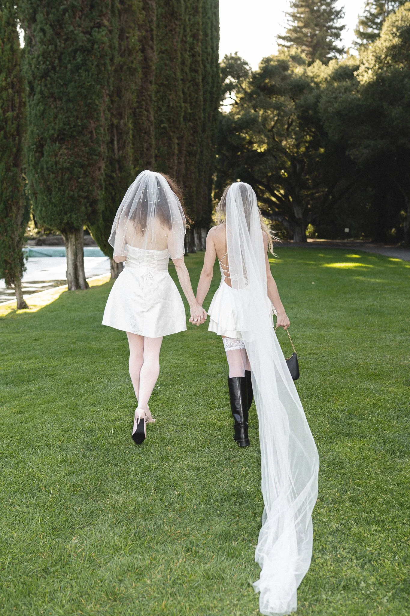 Two women dressed in white wedding dresses holding hands and walking away on a grassy area with tall trees in the background near Pulgas Water Temple in Woodside. Same sex couple photography.
