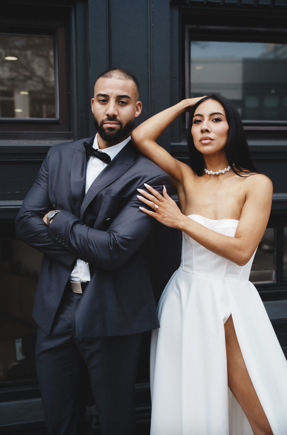 A man and woman dressed in formal attire posing together outside near a dark building with windows, elopement in North Beach neighborhood in San Francisco, California.