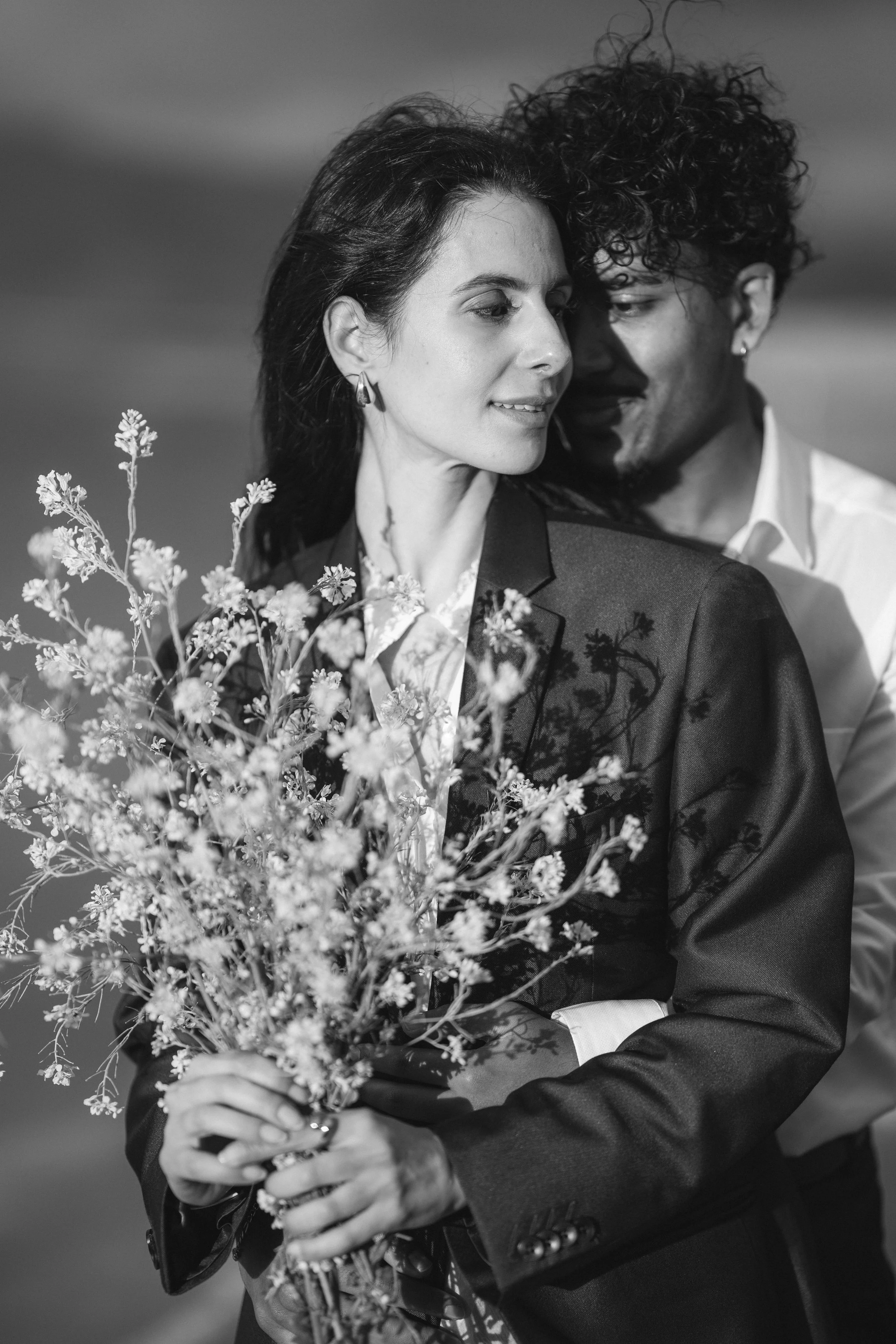 A man and woman standing closely, with the woman holding a bouquet of flowers. They appear to be sharing an intimate moment, smiling softly at each other, near pink lake in Marina Alviso.