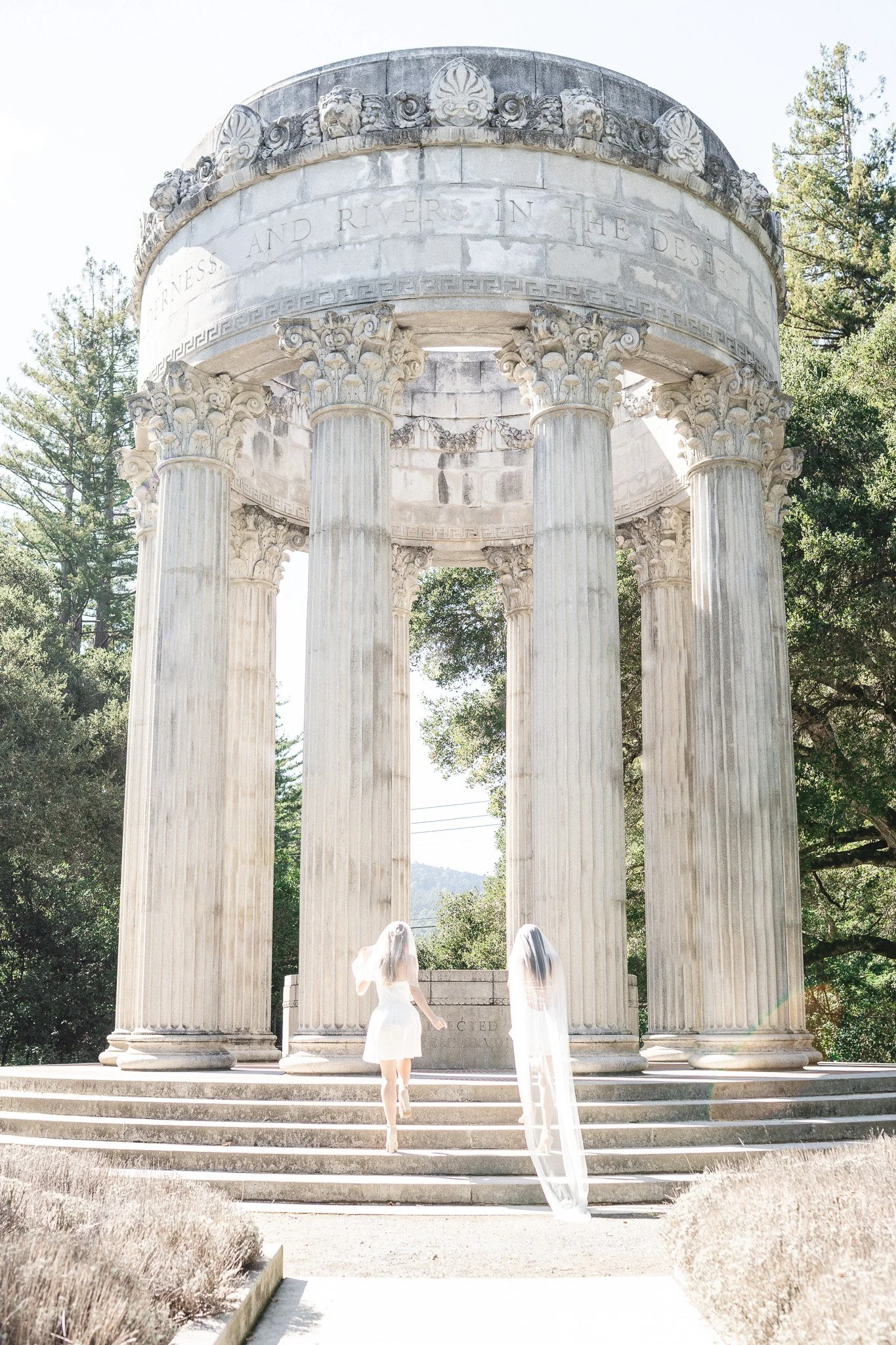 Two brides in white dresses with veils are descending the steps in front of the monument. Lesbian photography in Pulgas Water Temple, Woodside.