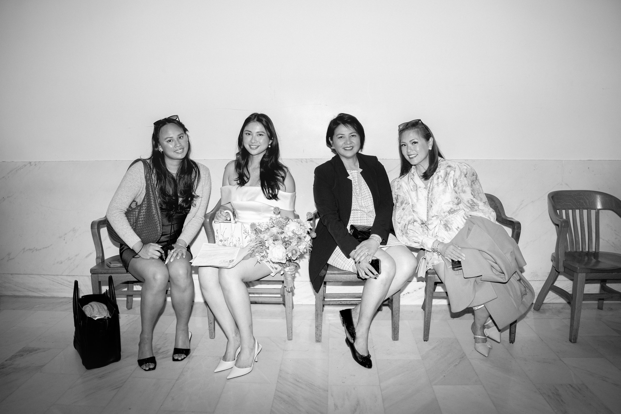 Four women sitting on a bench against a plain wall, smiling for the photo, with handbags and a bouquet on the bench inside San Francisco City Hall.