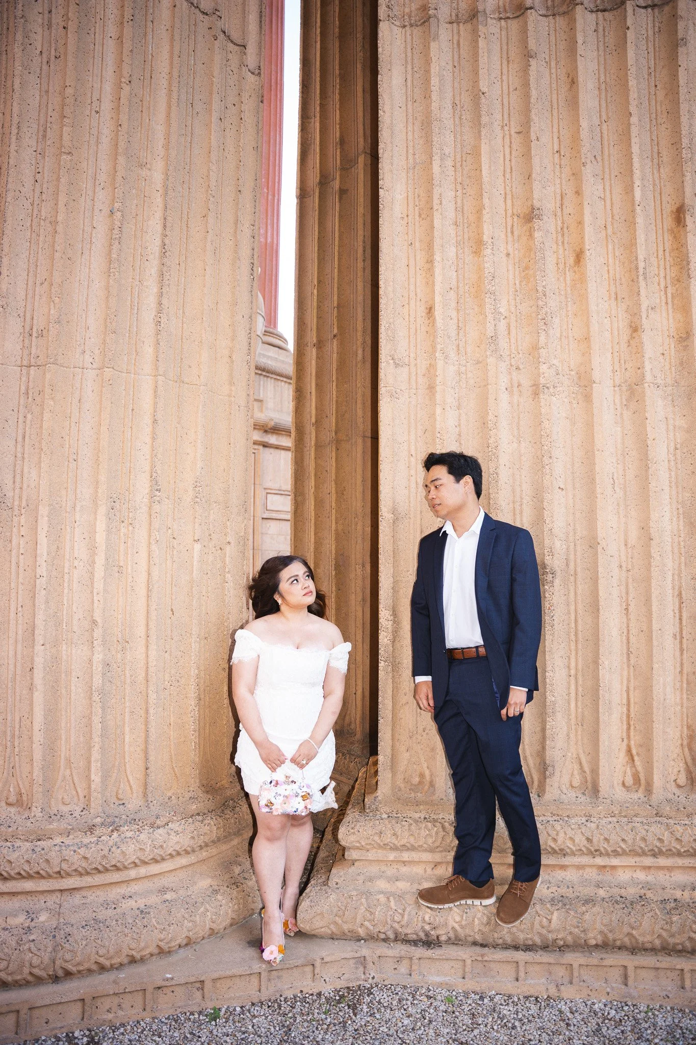 A woman in a white dress holding a bouquet and a man in a blue suit and brown shoes at the Palace of Fine Arts in Marina District of San Francisco.