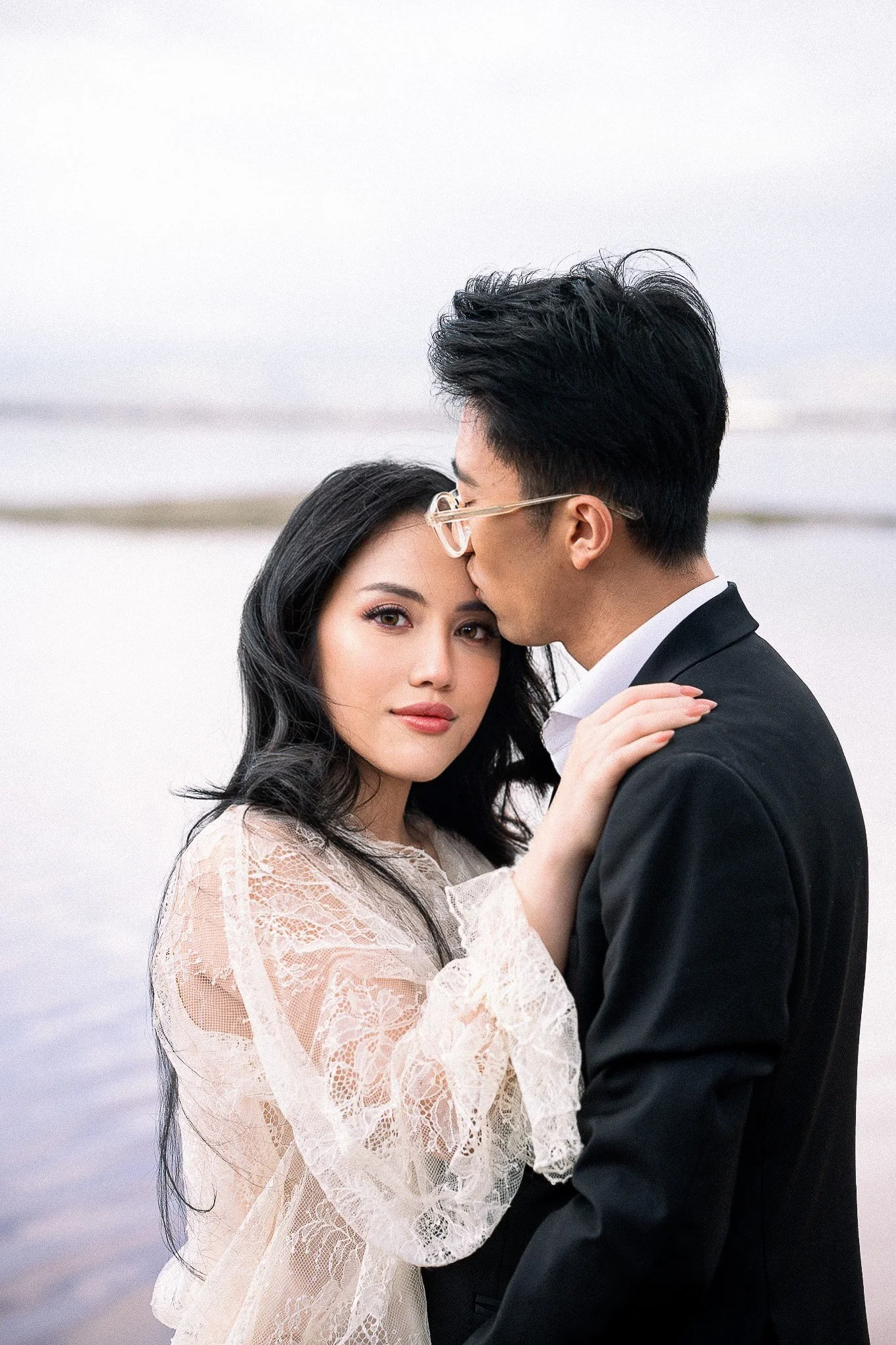 A bride and groom embrace at Marina Alviso pink lake, with the groom kissing the bride on the forehead. The bride has long black hair and wears a lace wedding dress, while the groom wears glasses and a black tuxedo.