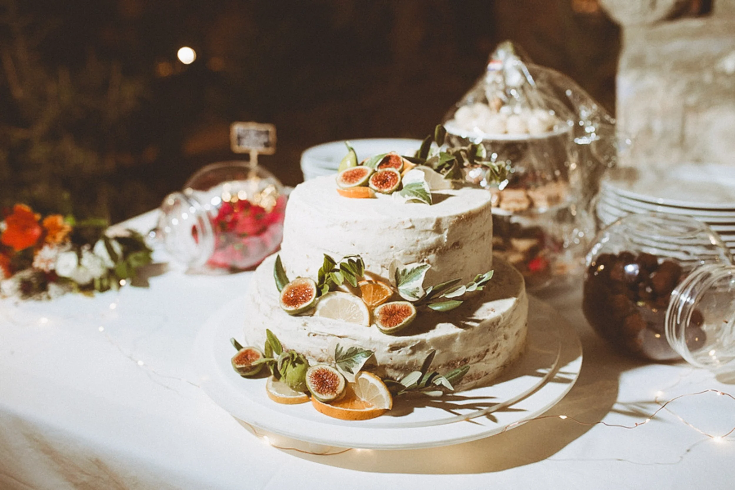 A layered cake decorated with sliced figs, oranges, and green leaves on a white serving platter, with additional desserts and plates in the background. Flash night photography.