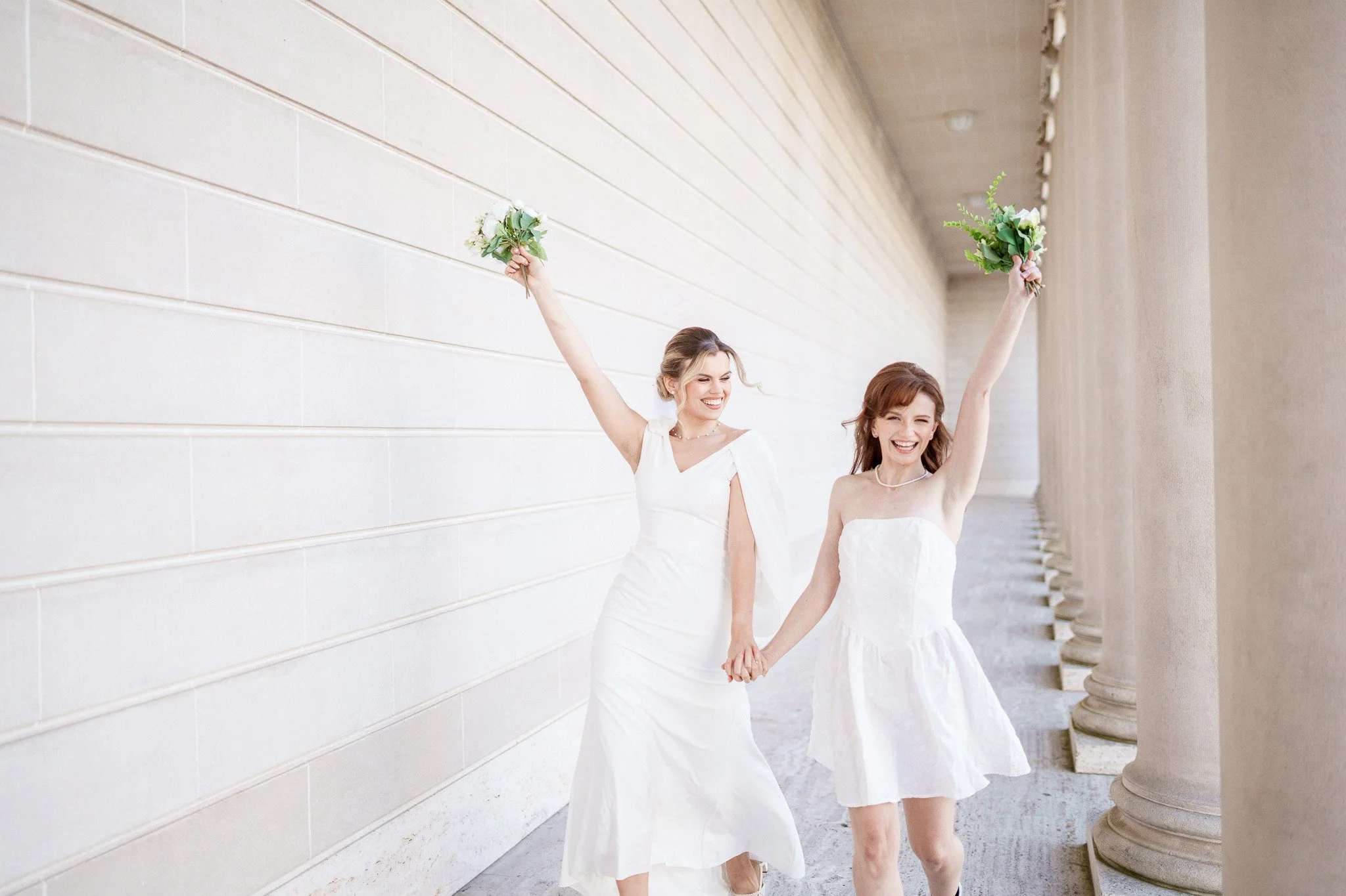 Two women in wedding dresses holding hands and bouquets, standing near columns in an outdoor corridor with a cream-colored wall. LGBTQ+ wedding, Legion of Honor museum.